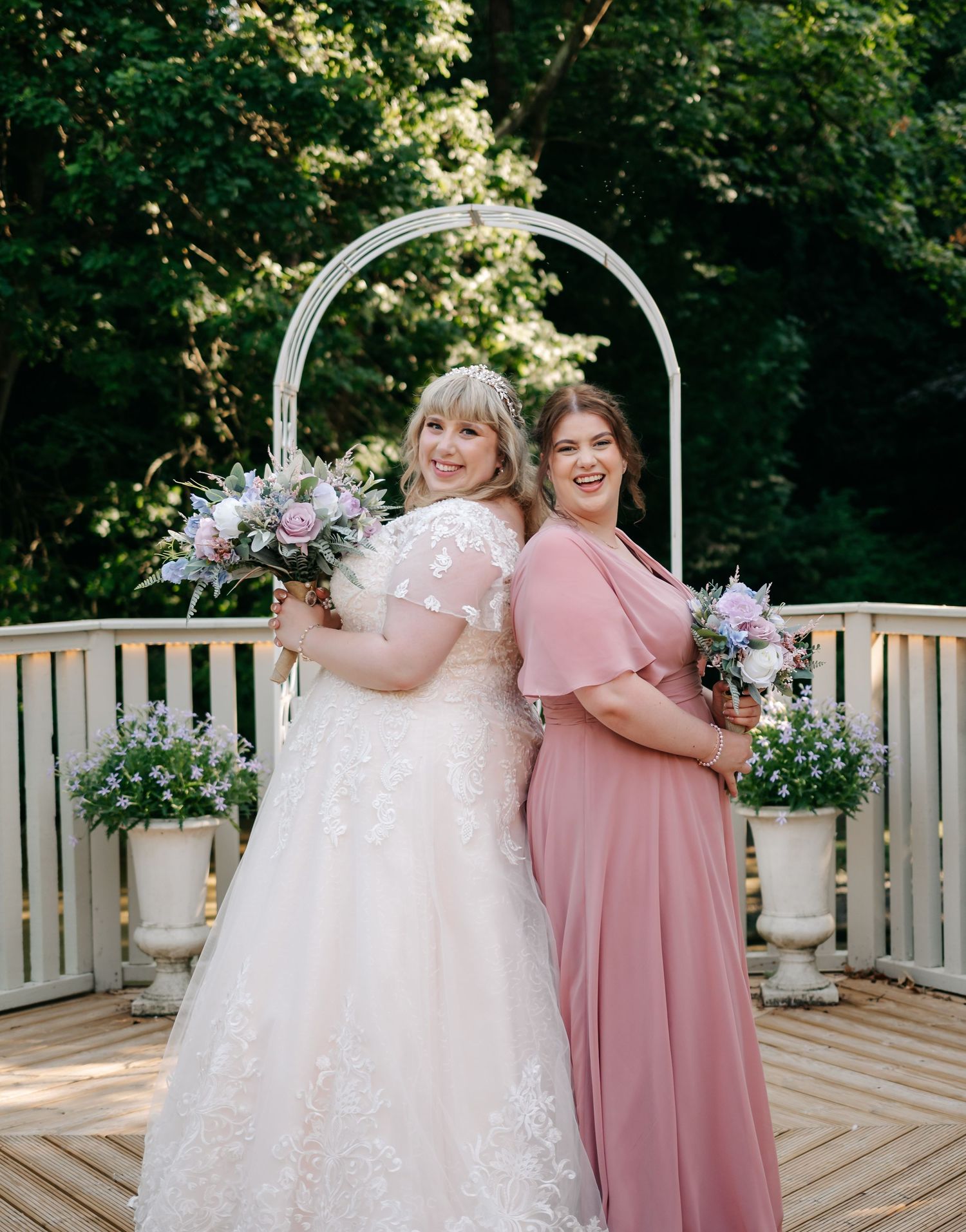 Bride in lace wedding gown and bridesmaid in dusty rose dress posing before white arch outdoors.