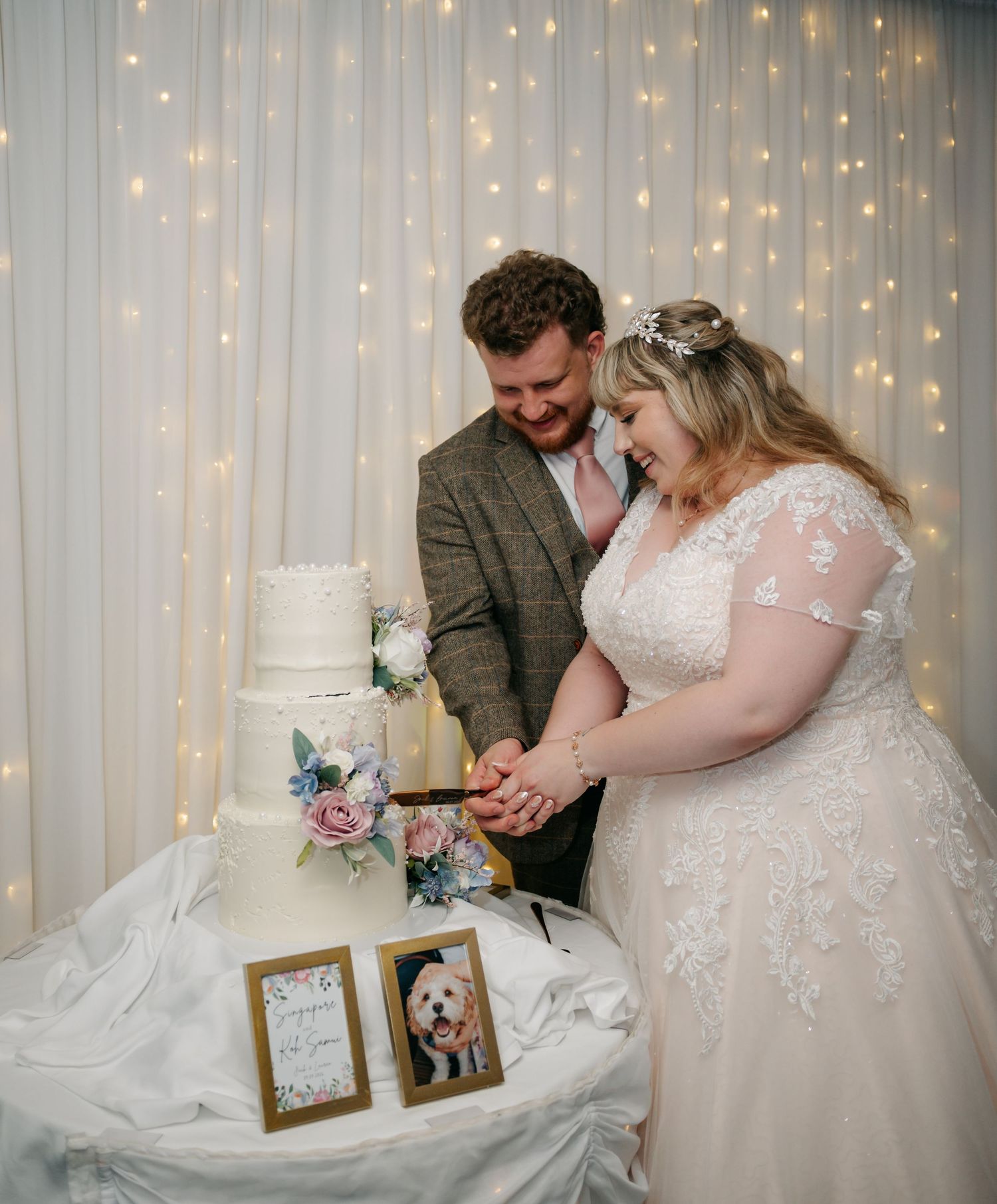 Bride and groom cut their elegant white tiered wedding cake in front of a fairy light backdrop.