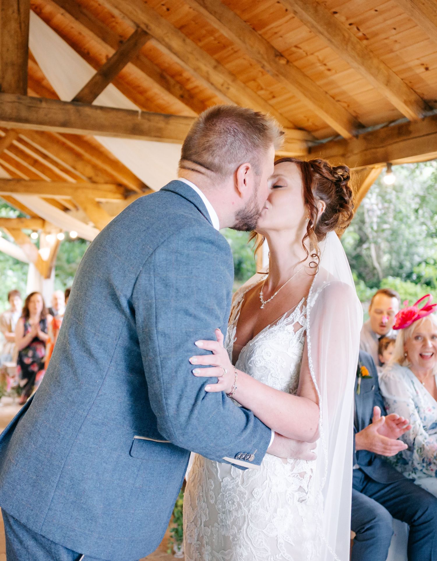 Romantic wedding kiss under a wooden pavilion surrounded by guests.