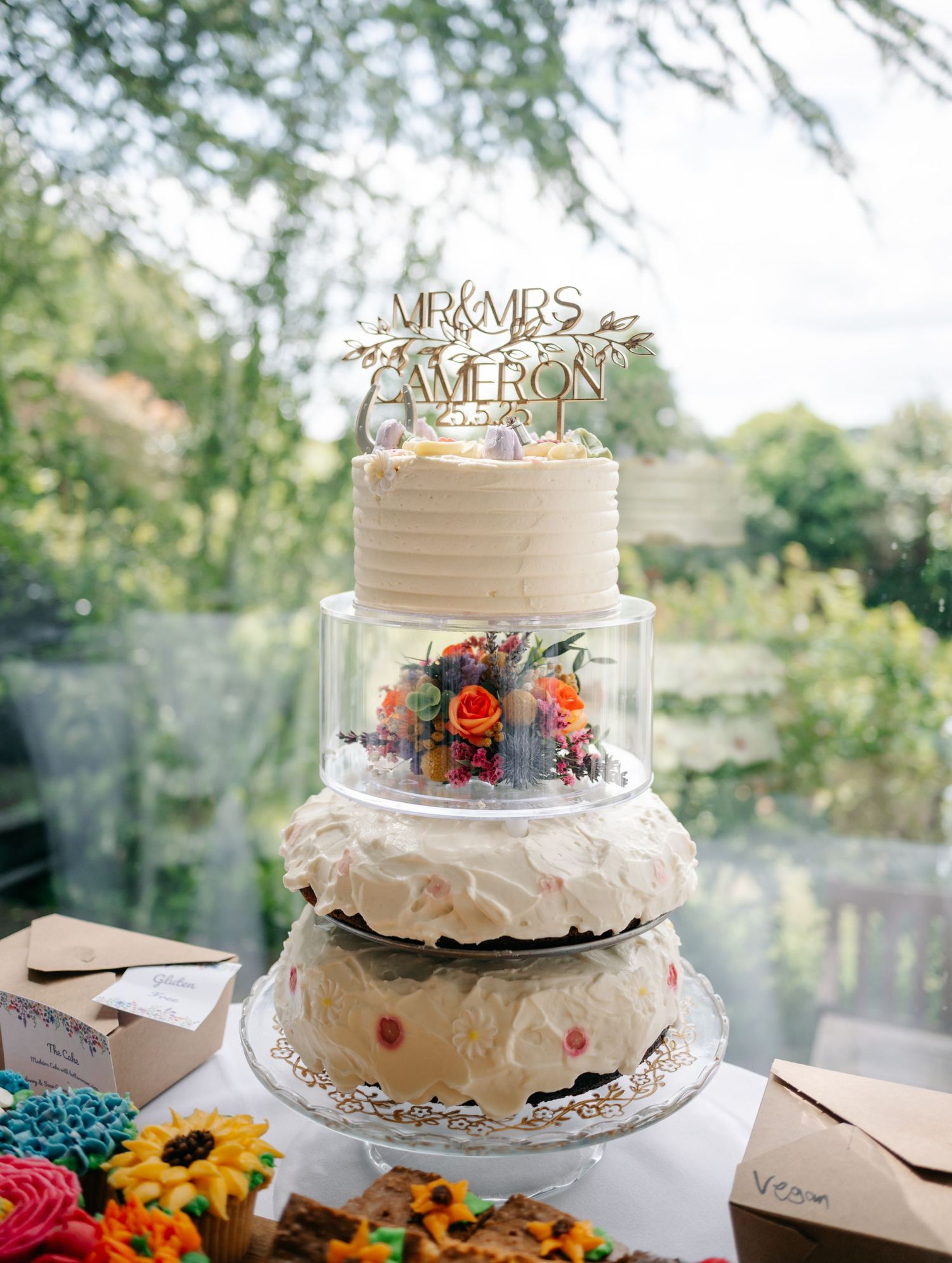 Two-tiered white wedding cake decorated with floral accents and a personalized topper.