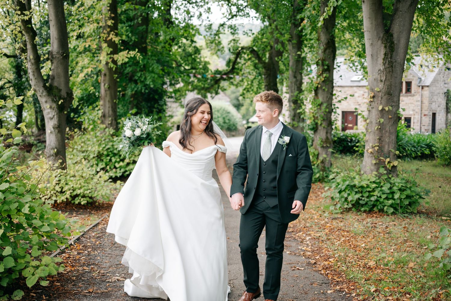 Newlyweds walk together through a lush green garden path surrounded by trees on their wedding day.