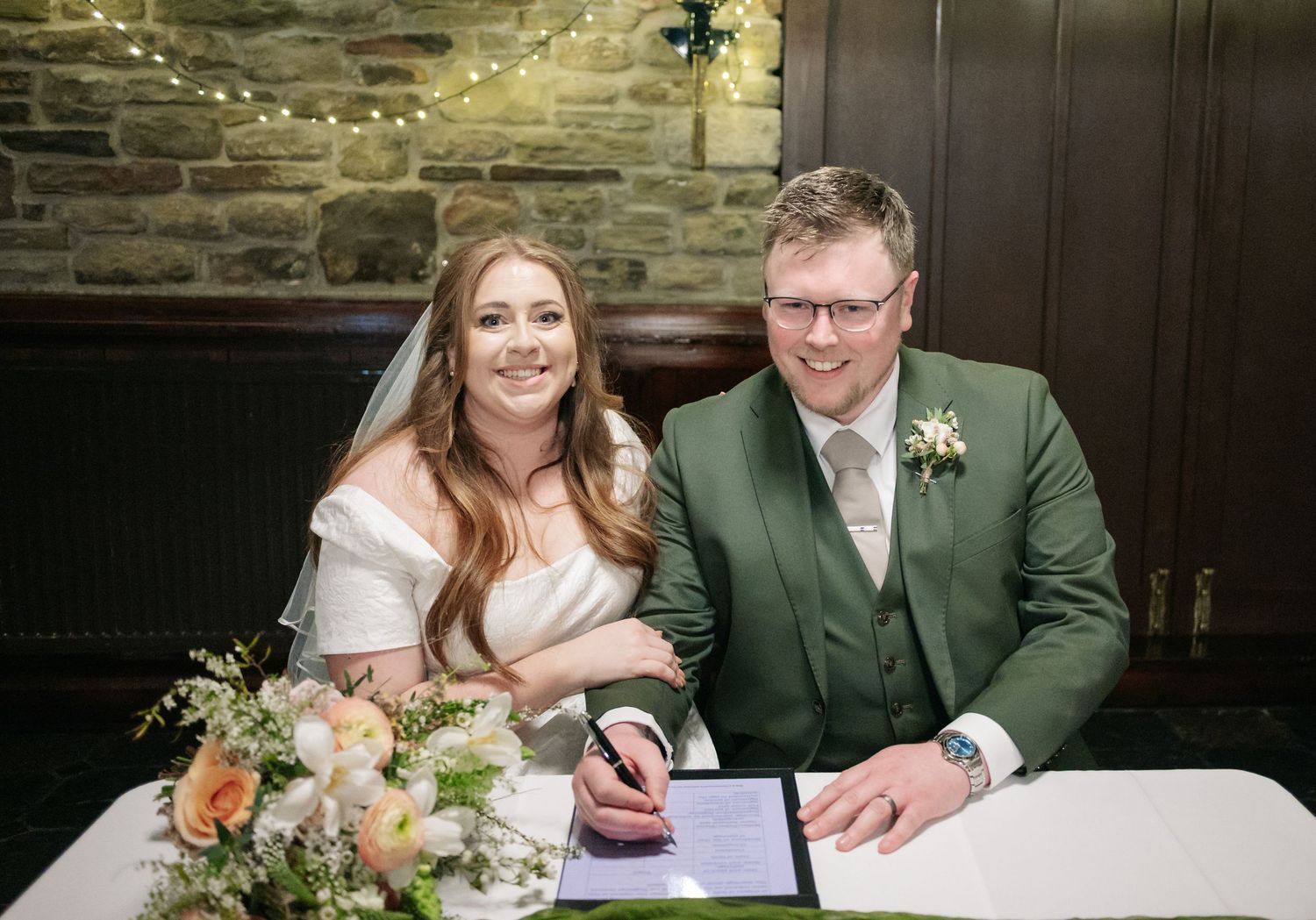 Wedding couple signing marriage documents at a wooden table in a rustic stone venue.