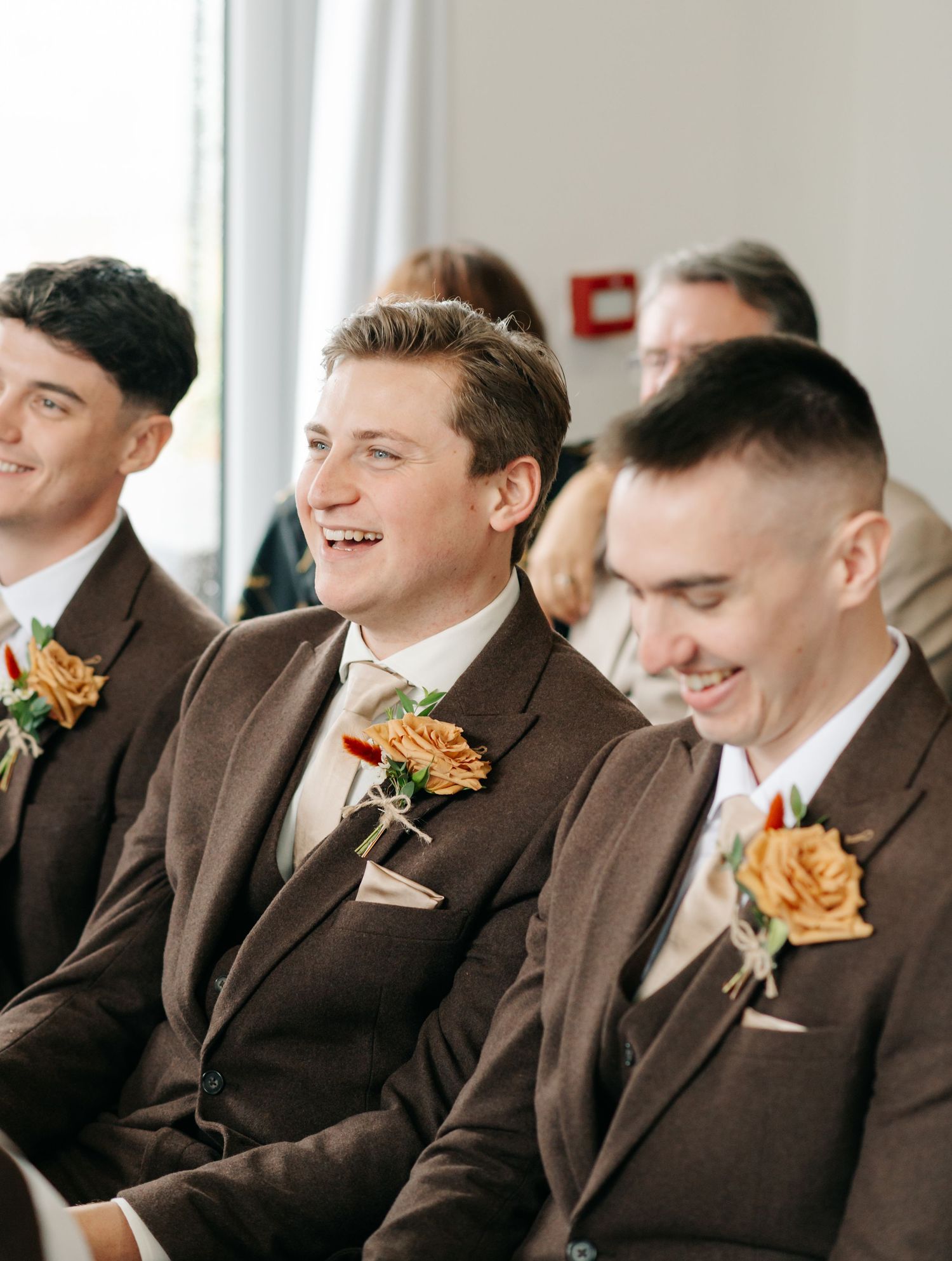 Groomsmen in matching brown suits with orange boutonnieres share a candid laughing moment at a wedding.