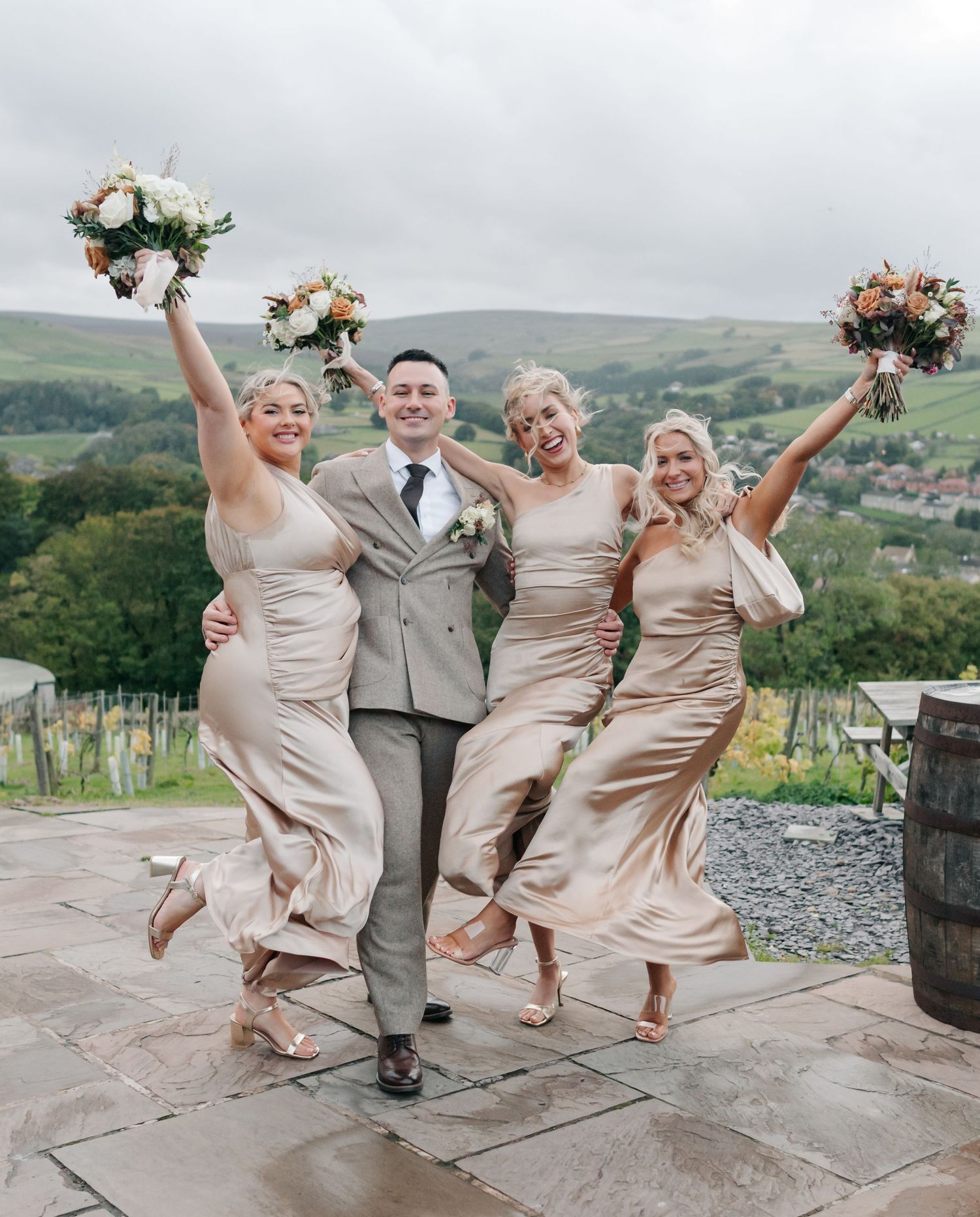 Wedding party jumping for joy on stone patio with scenic countryside views in background.
