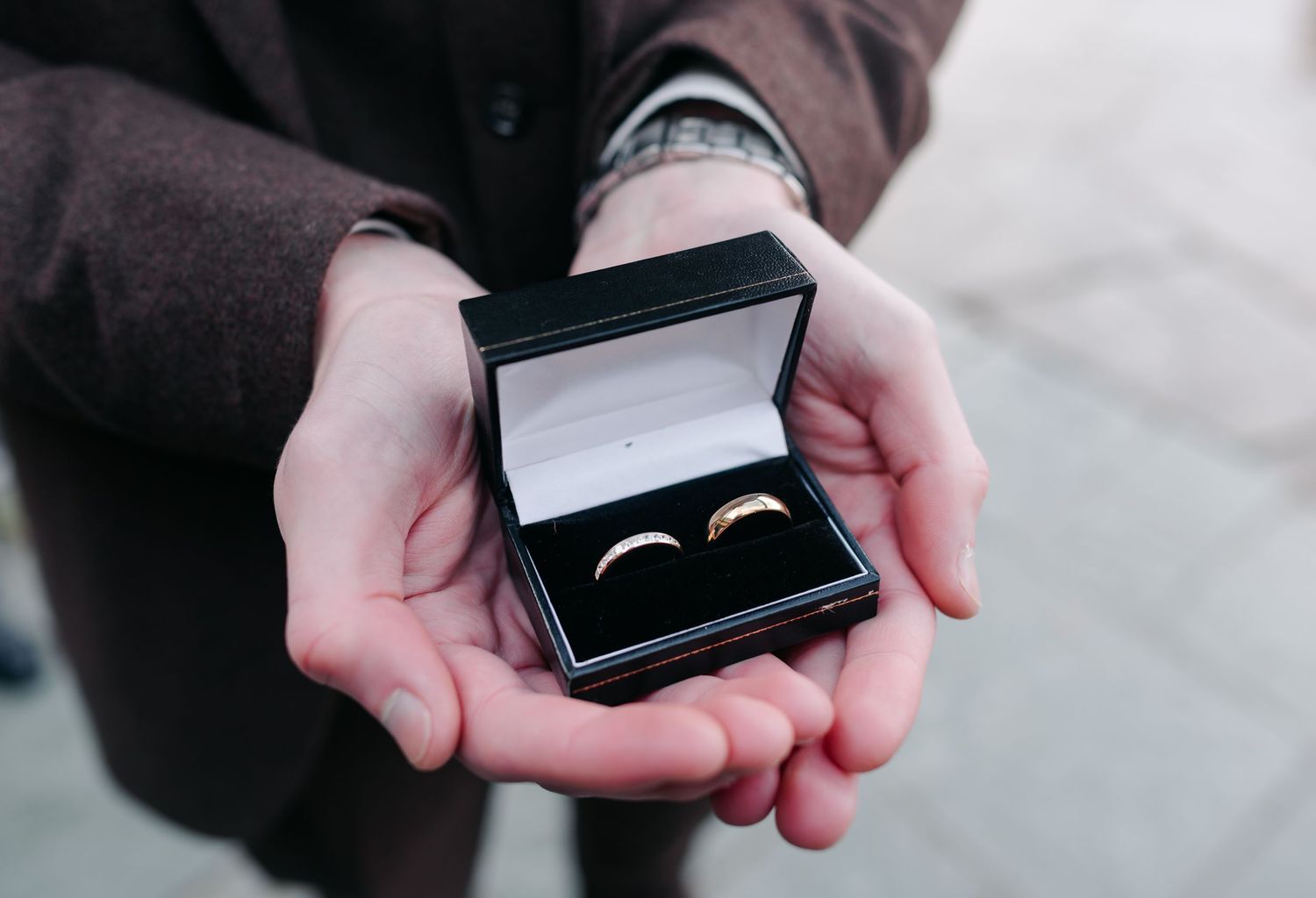 Hands holding an open black jewelry box containing two gold wedding rings against a blurred background.