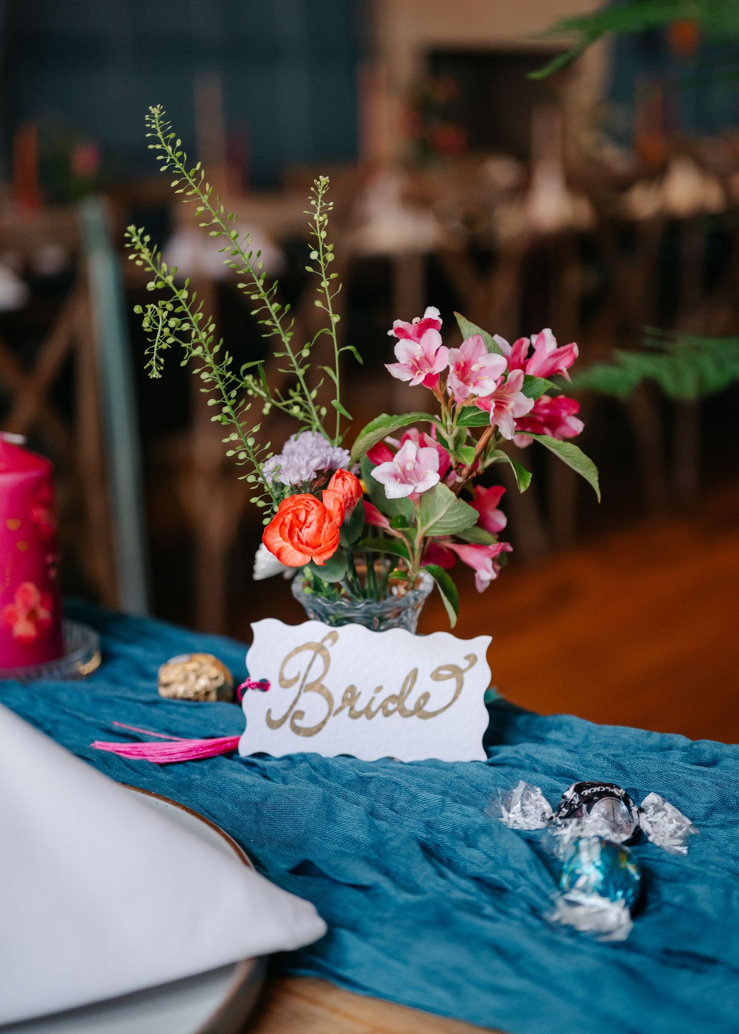 Wedding table setting with pink flowers in vase, blue tablecloth, and elegant 'Bride' place card.