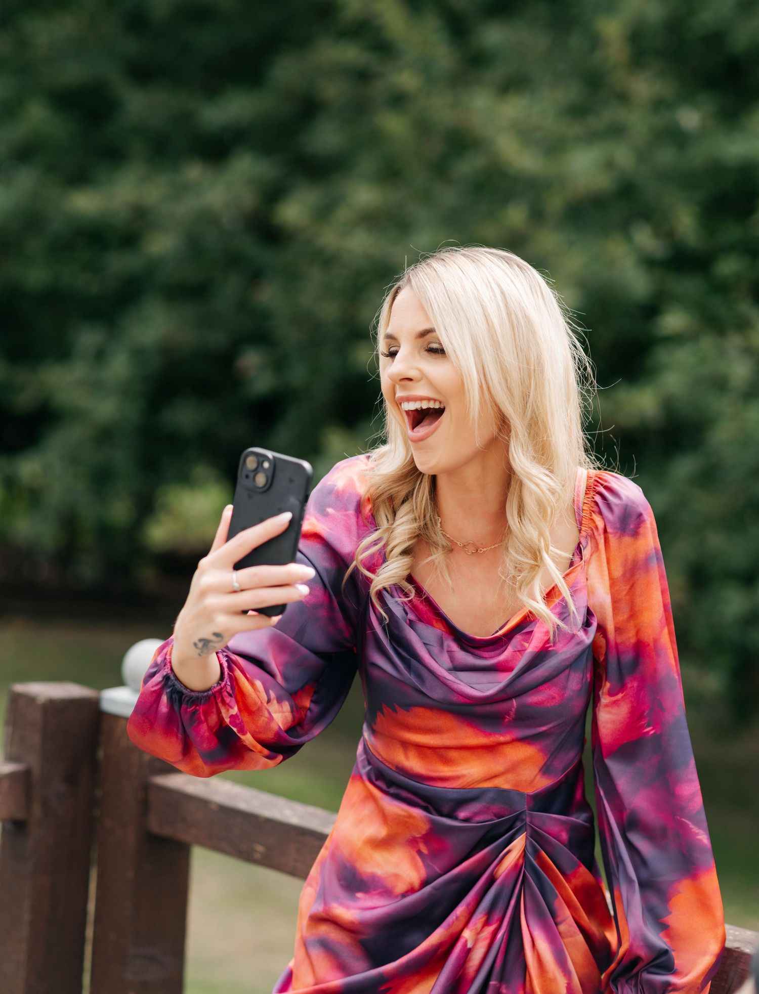 Woman in vibrant tie-dye dress poses playfully with phone during outdoor photoshoot.