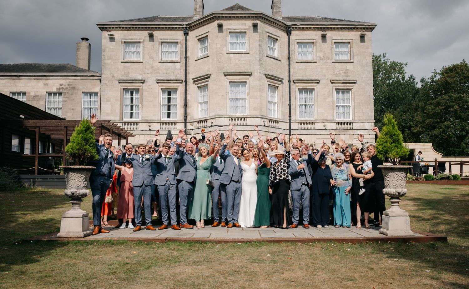 Large group wedding photo of guests and wedding party in front of a grand estate building.