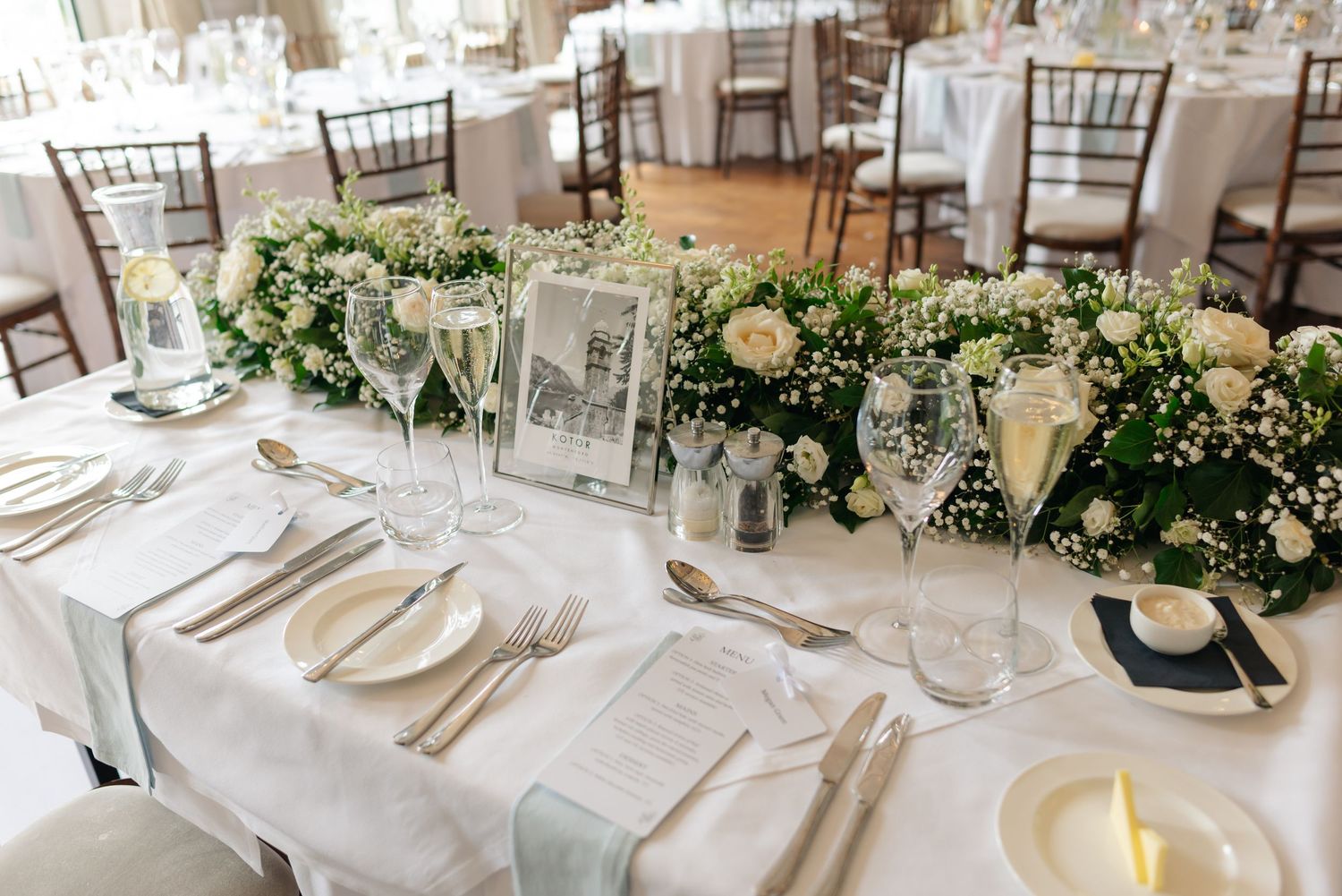 An elegant wedding reception table with white linens and floral garland centerpieces featuring vintage photo frames.