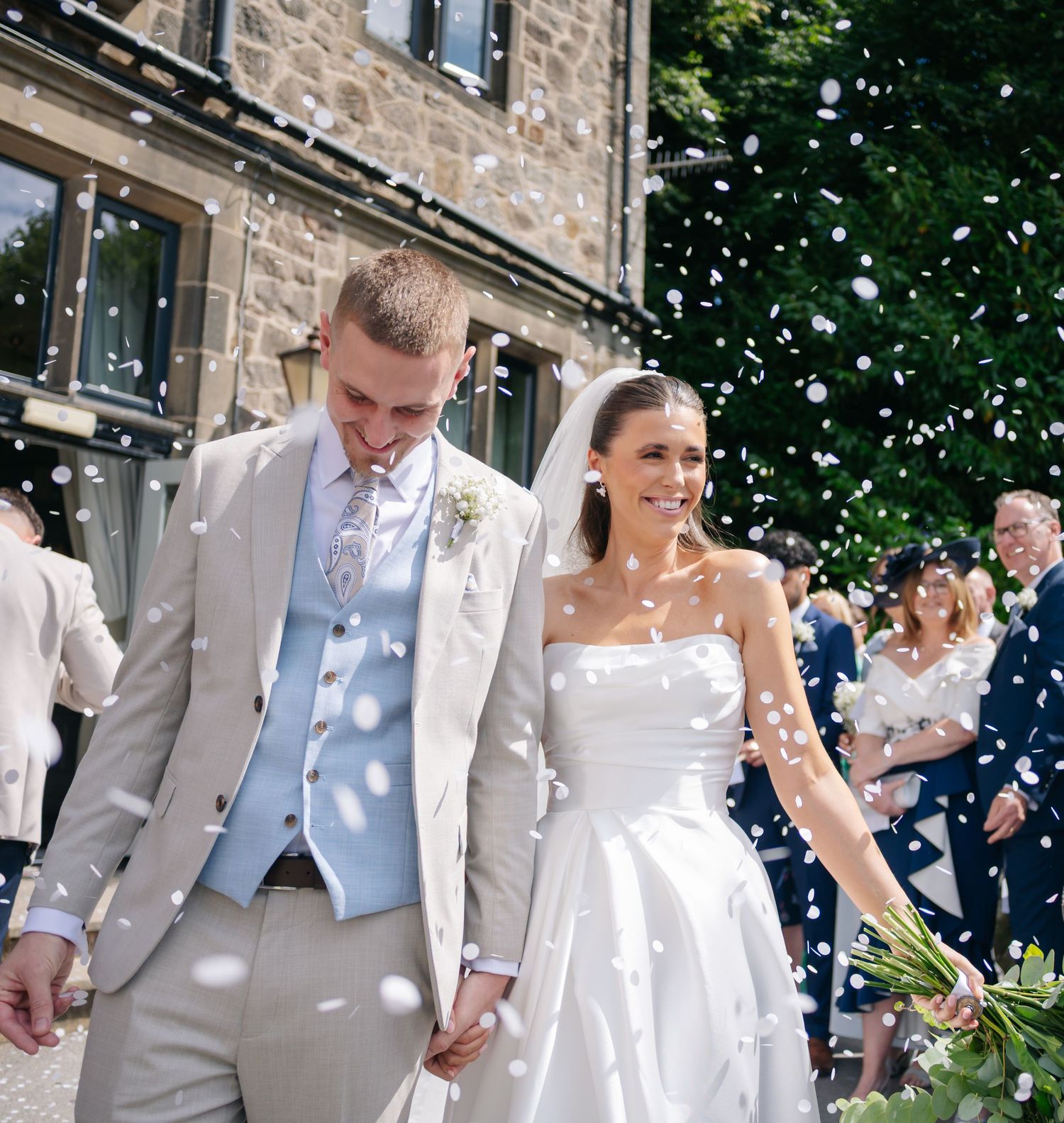 A couple walks hand in hand through falling confetti outside a stone building.