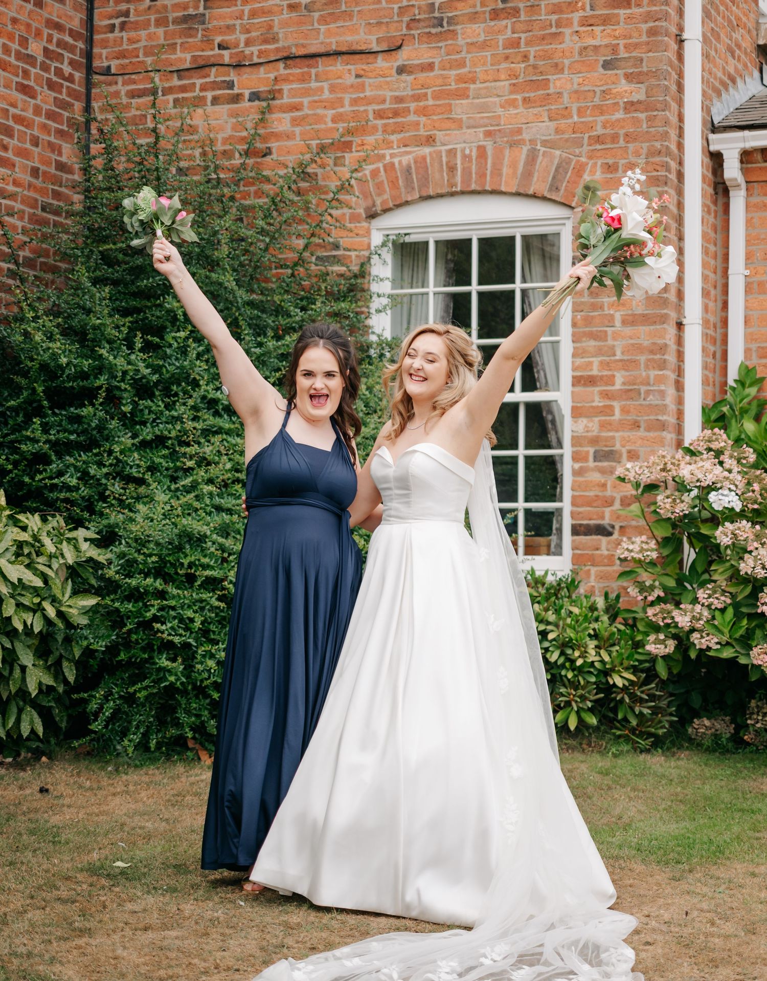 Two people celebrate joyfully outside a brick building, one in navy and one in a white wedding gown.