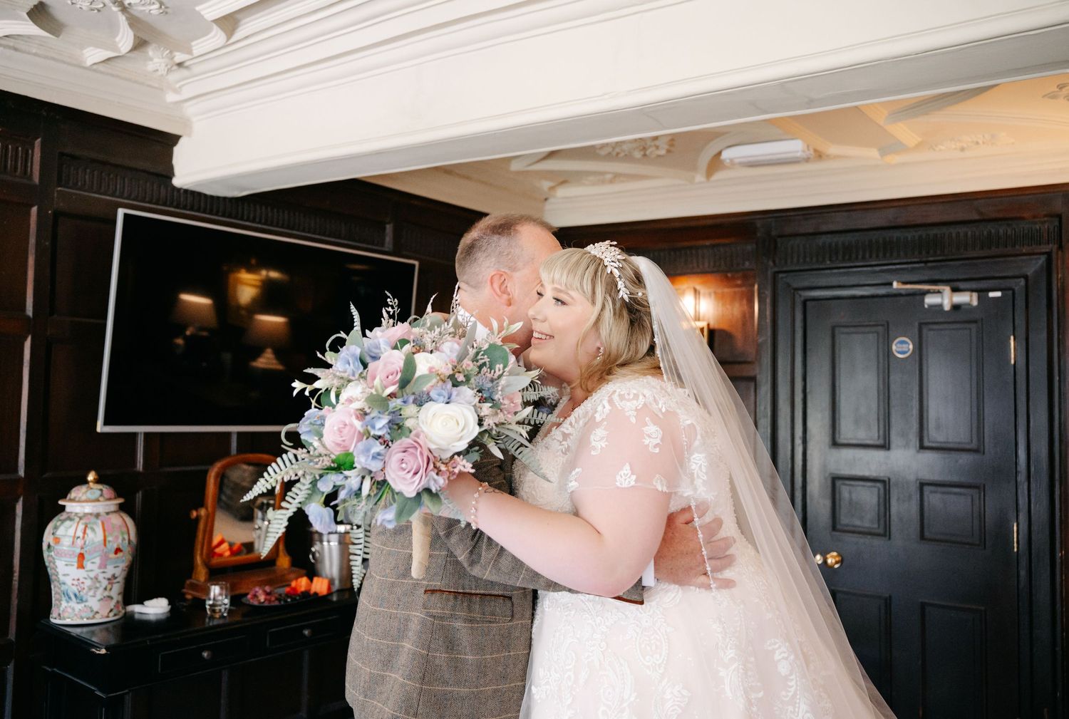 Bride and groom share a tender kiss, bride holding a pastel bouquet in an elegant dark-wood venue.