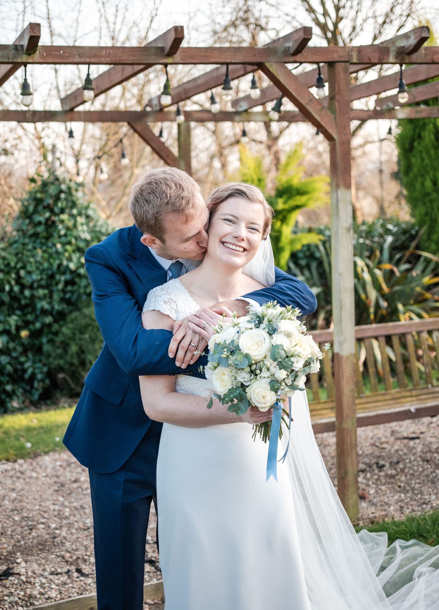 Wedding couple embracing under a wooden pergola with white floral bouquet and navy suit.
