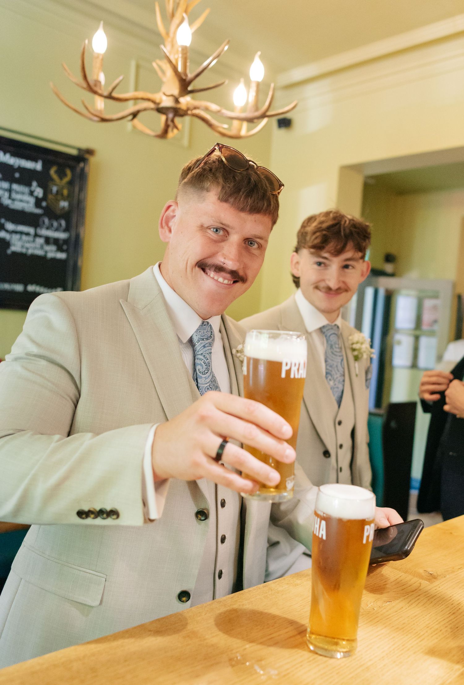 Wedding party members in beige suits share beers and laughs at a wooden bar counter.