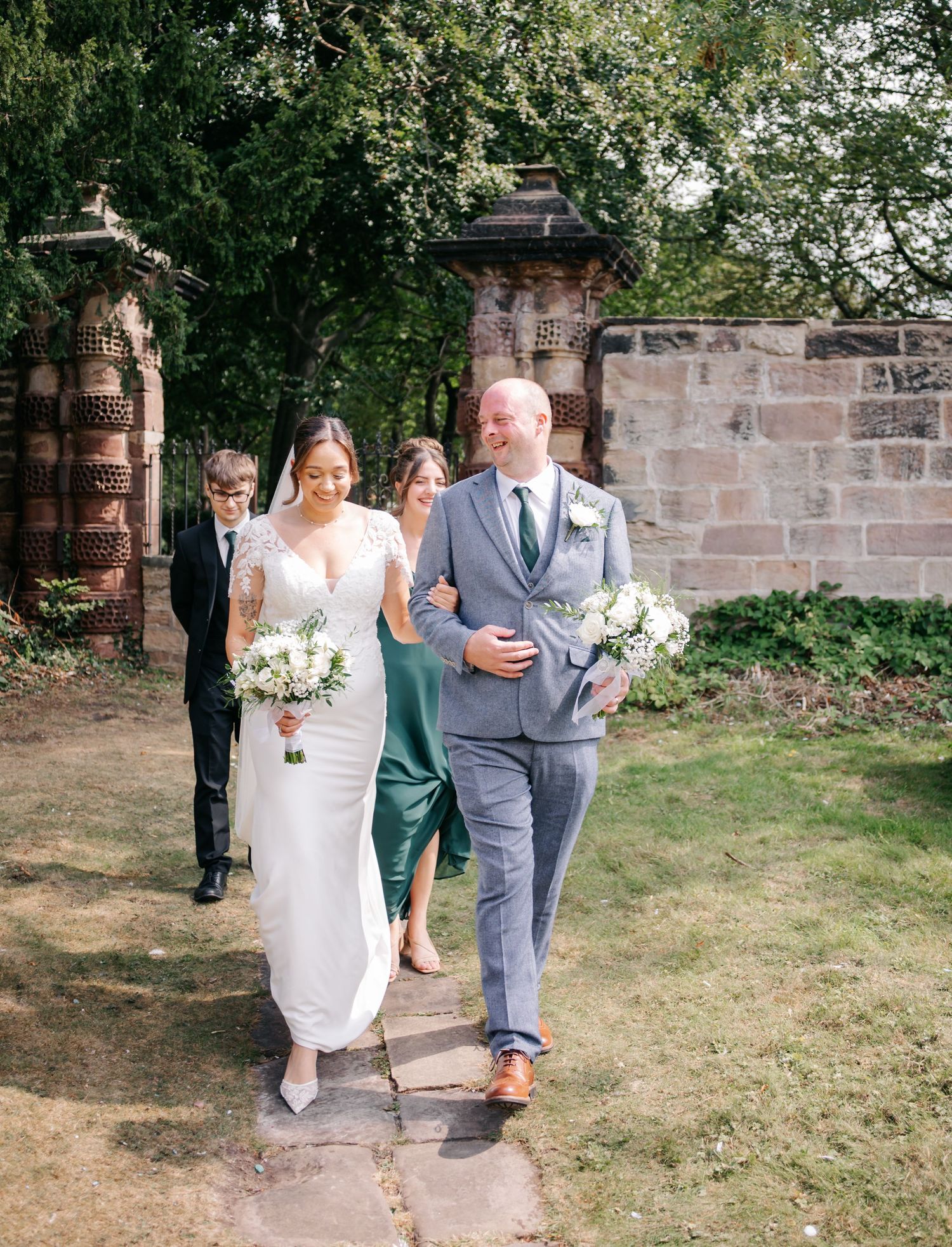 Wedding party members walk along a garden path during an outdoor celebration.