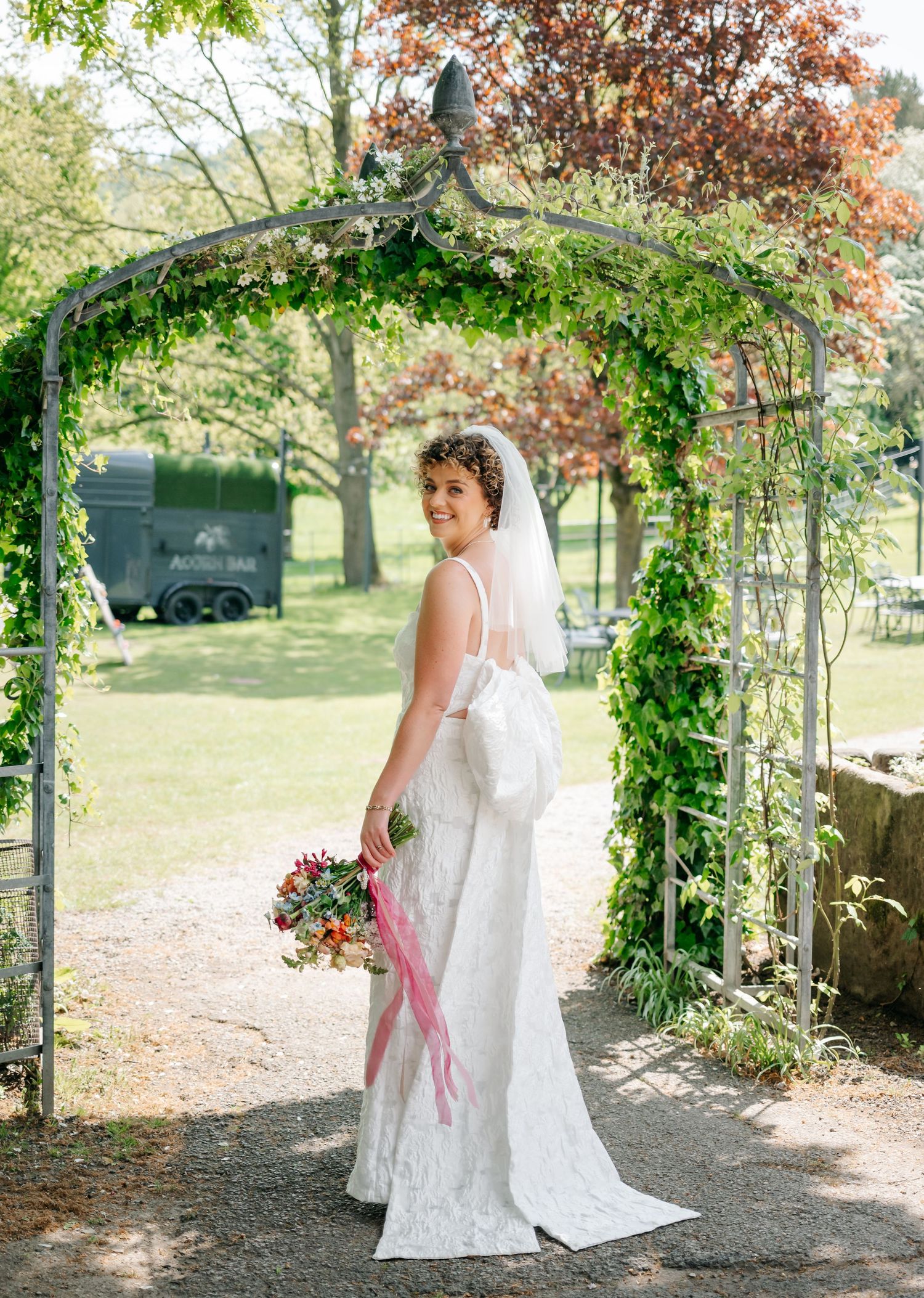 A wedding dress hangs elegantly under a vine-covered garden archway with autumn trees in the background.