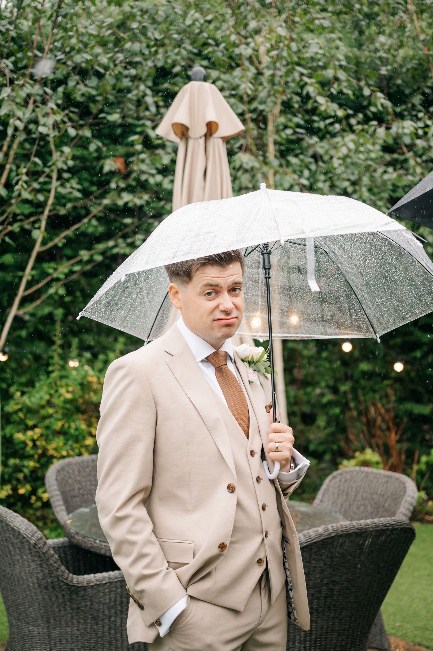 Wedding party members holding clear umbrellas in a garden setting wearing light beige suits.