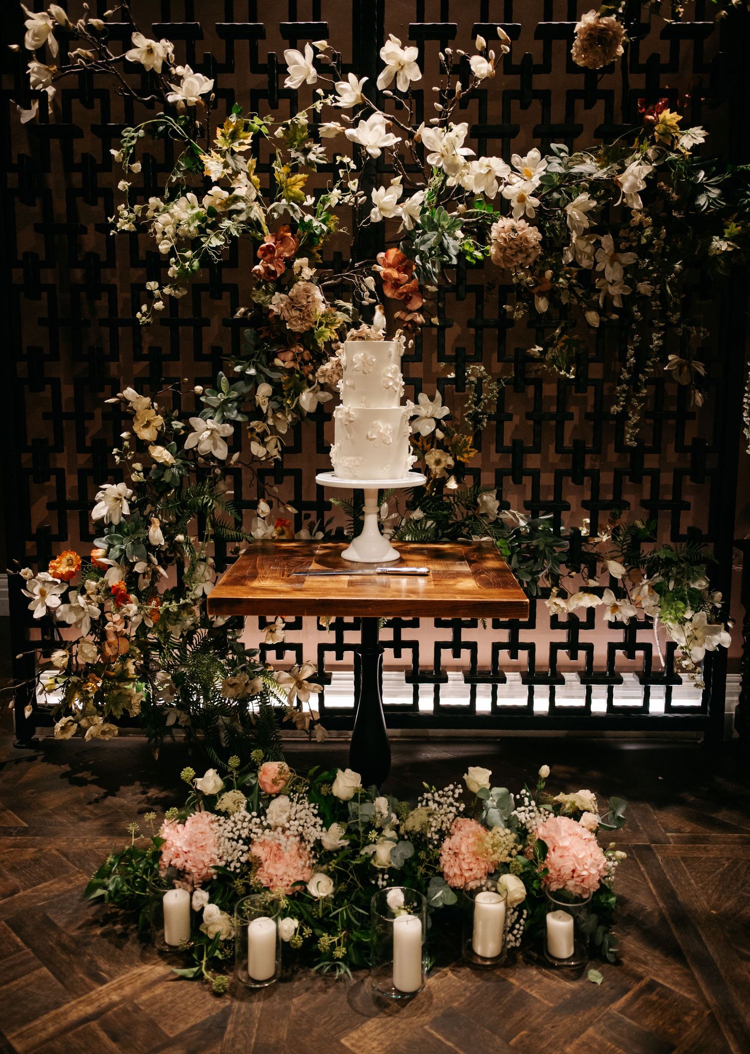 Elegant wedding cake display with lush floral arrangements and candles against a geometric backdrop.