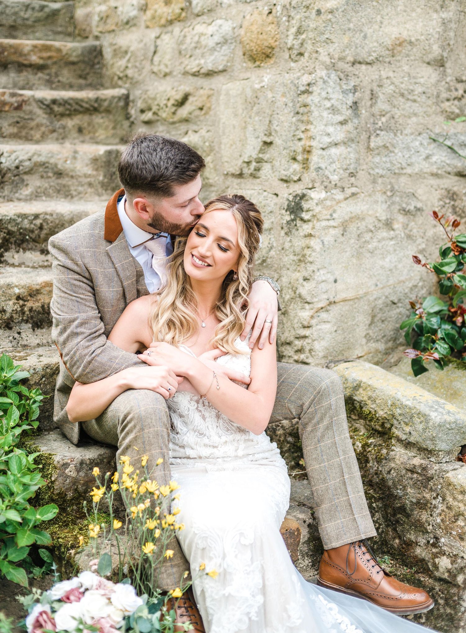Romantic couple sits together on weathered stone steps while embracing against rustic garden backdrop.