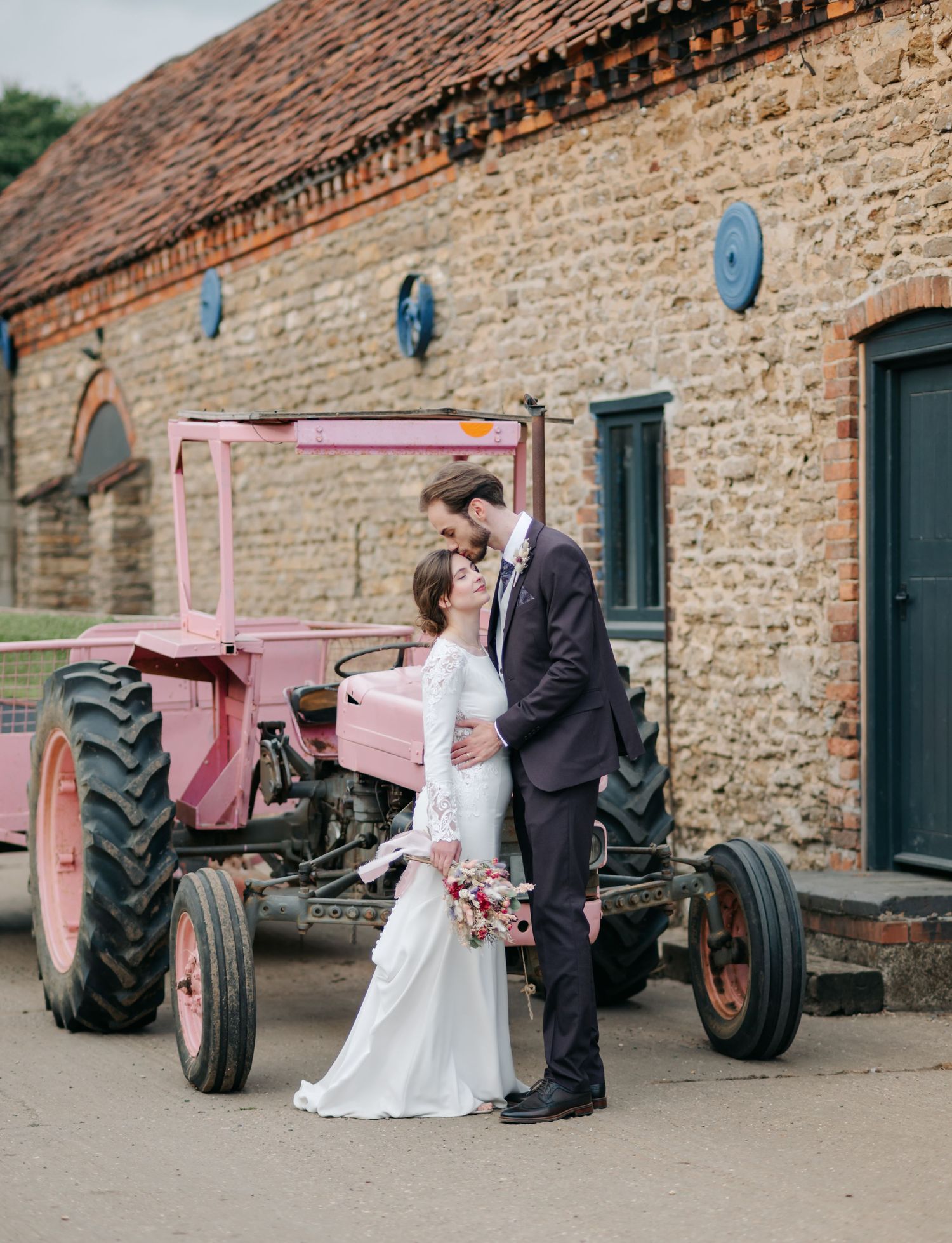 Wedding couple poses beside a vintage pink tractor against rustic stone building wall.
