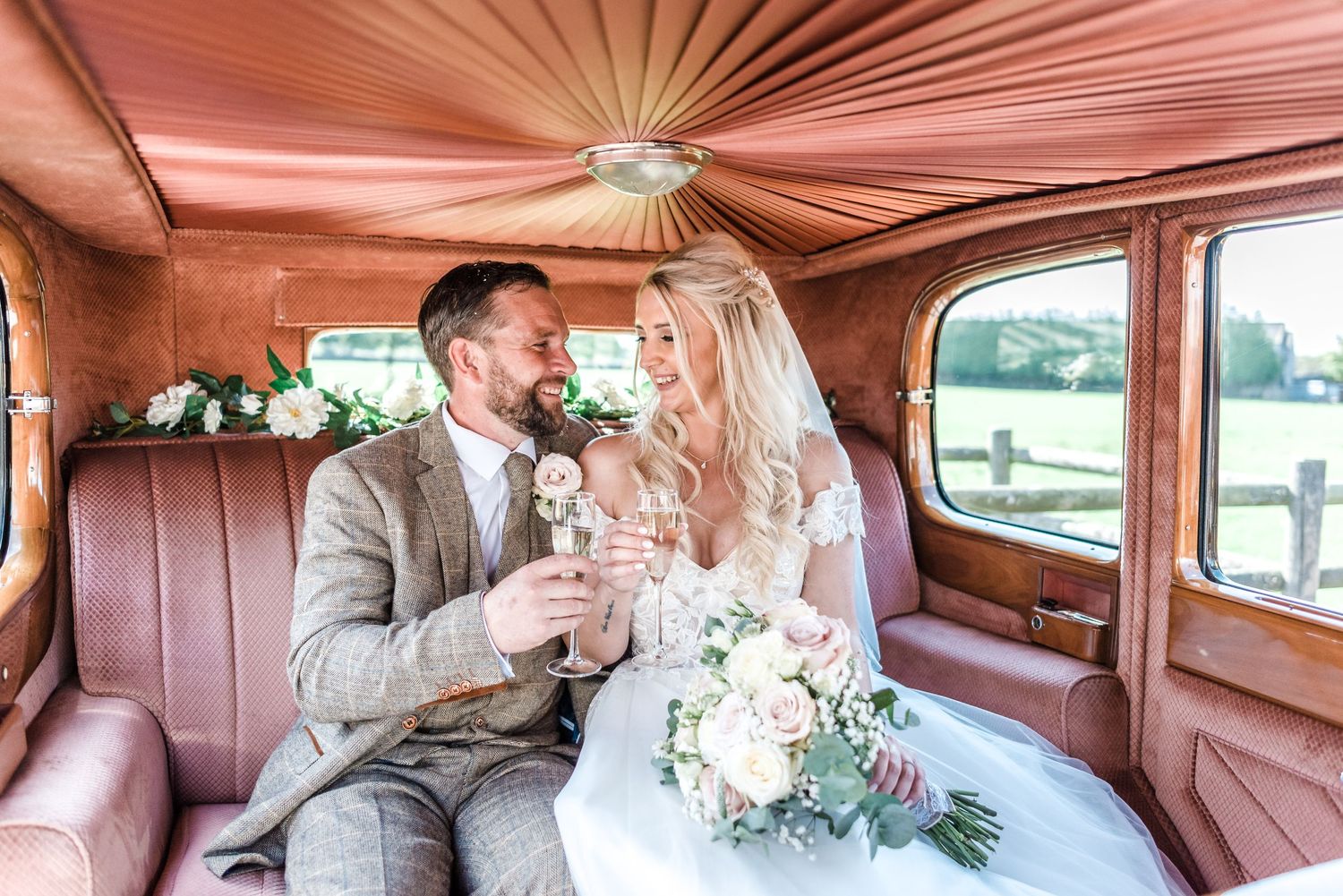 Wedding couple toasts with champagne inside classic car with pink interior.