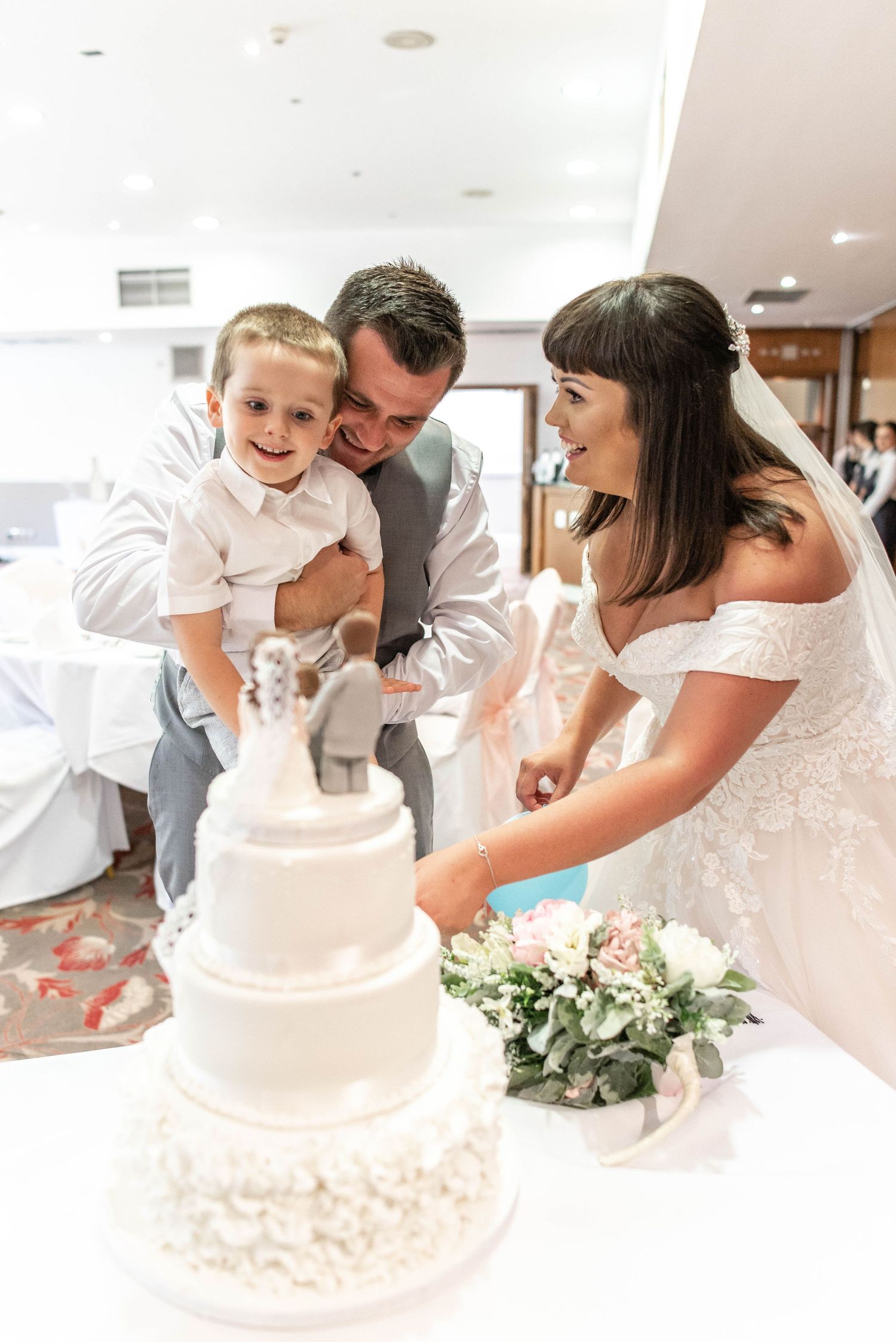 Wedding cake cutting moment in a bright reception hall with white tiered cake.