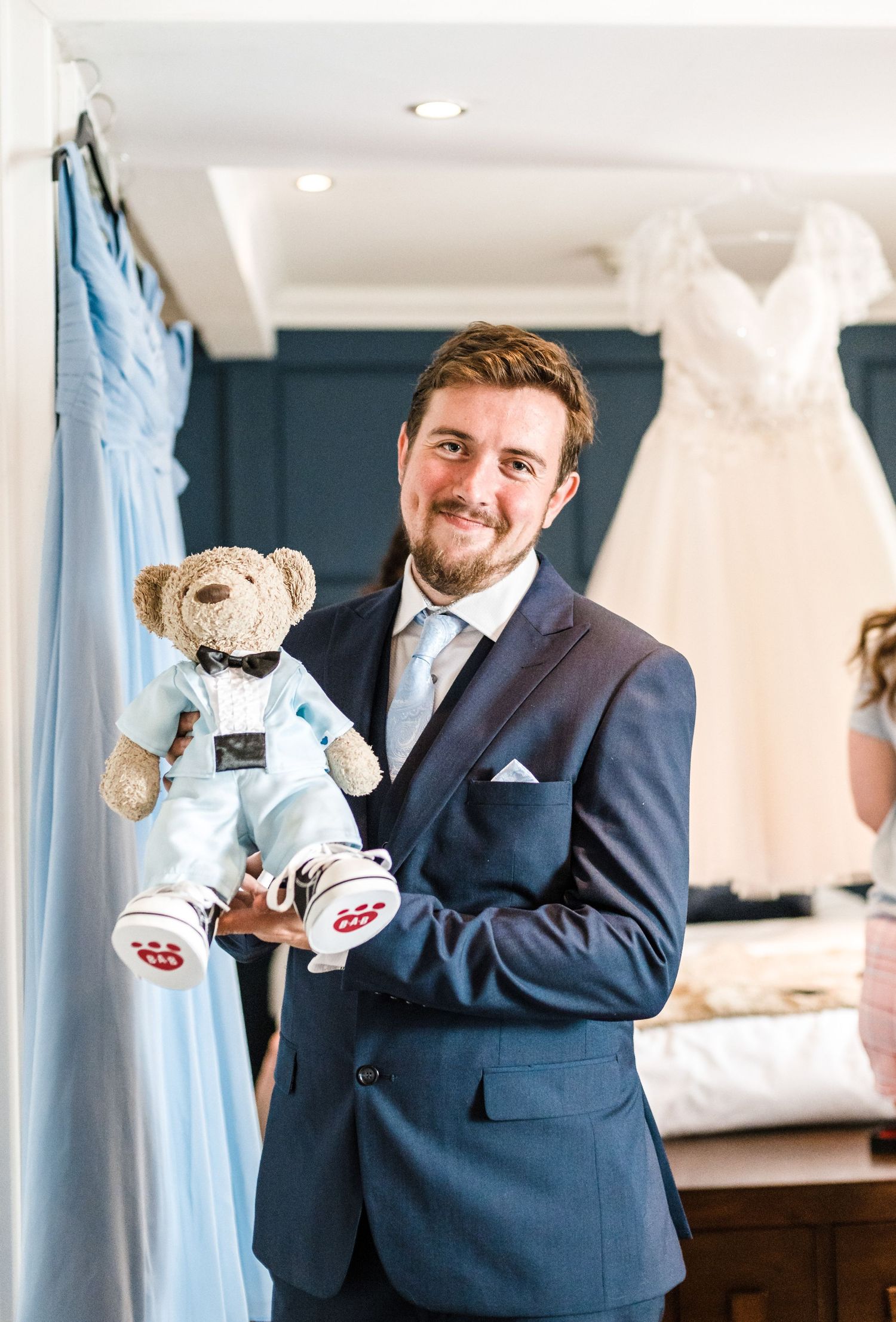 A formal-suited person holds a teddy bear wearing matching formal attire in front of wedding attire displays.