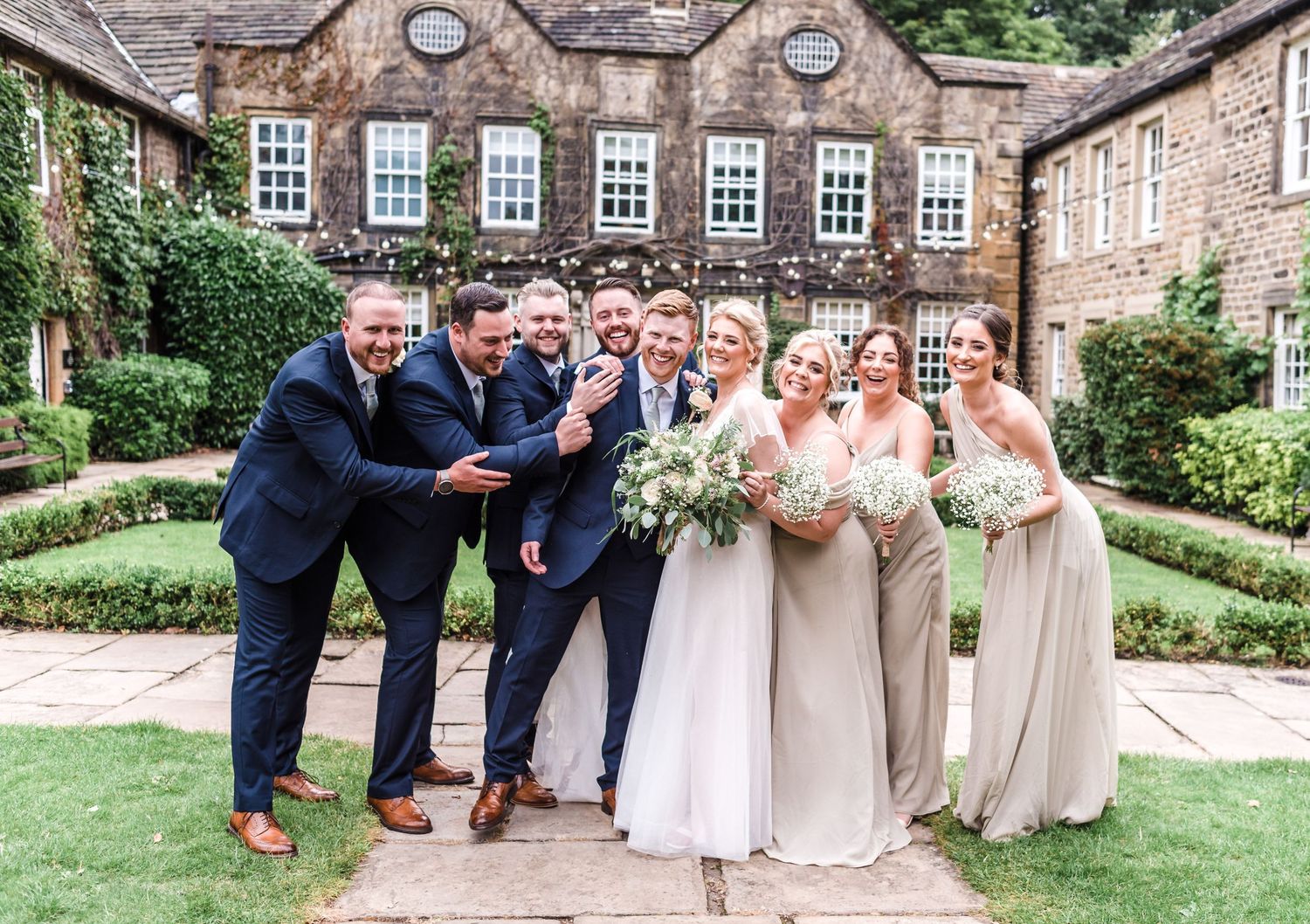 A wedding party poses for a fun group photo in front of a stone mansion with manicured gardens.