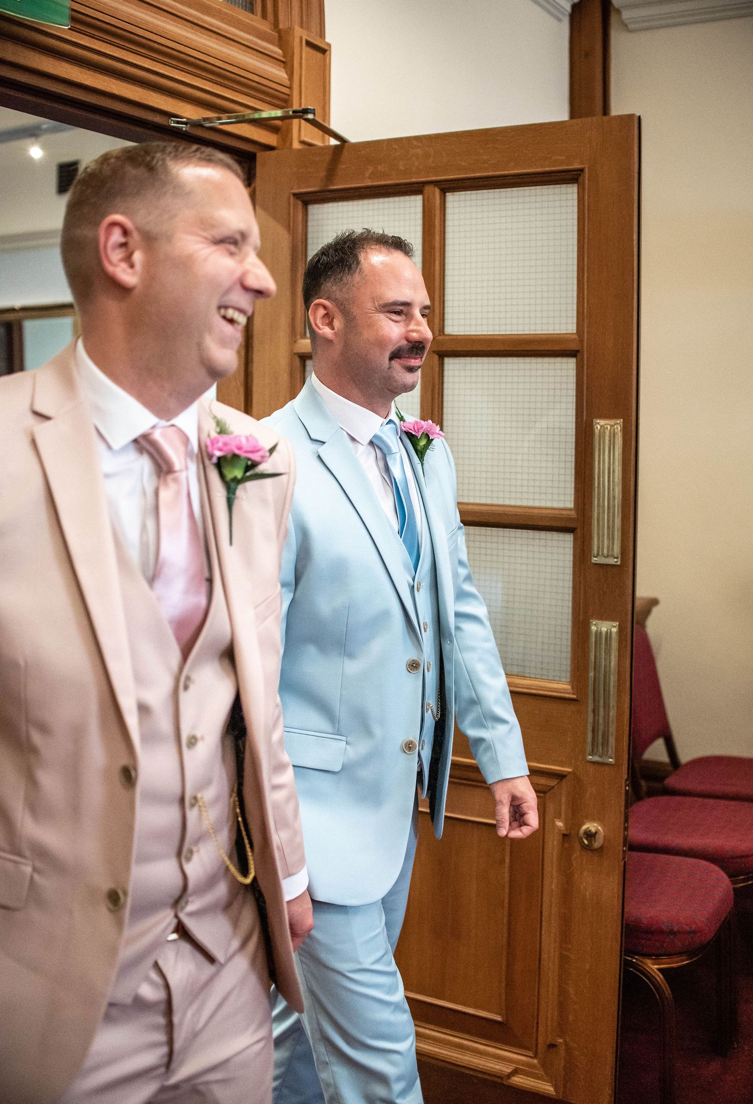 Two people in coordinating pastel suits share a joyful moment in a wooden-paneled hallway.