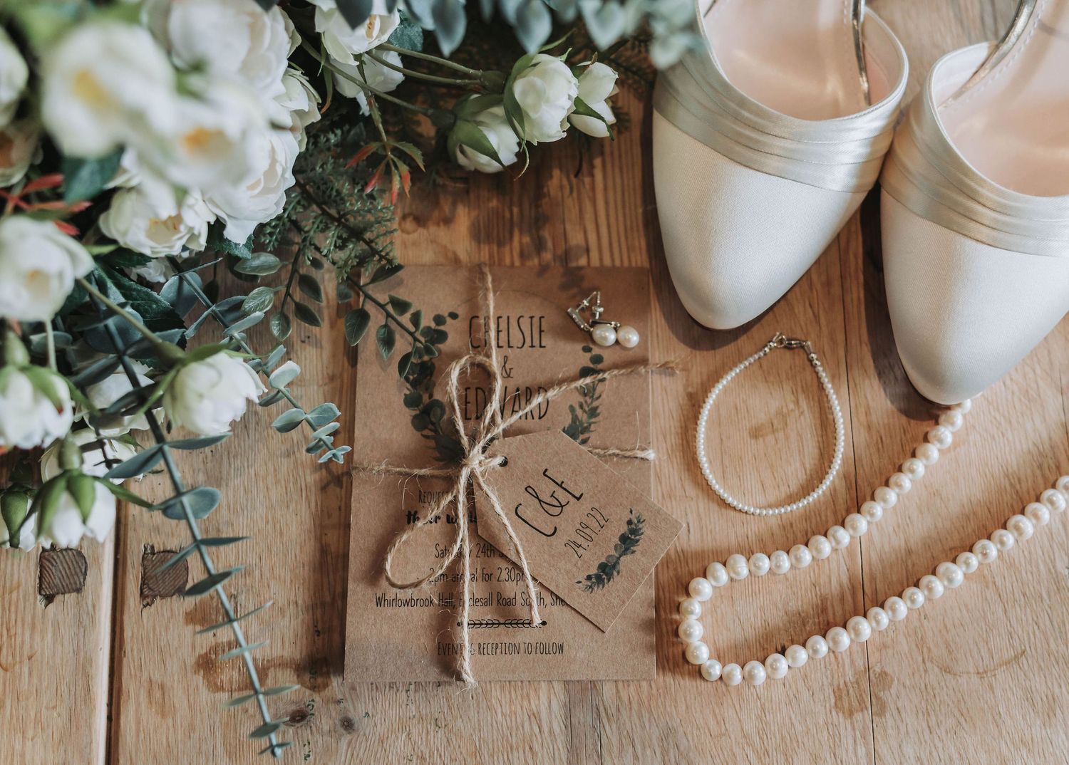 Wedding details flatlay with white shoes, pearls, flowers and vintage-style stationery on wooden surface.