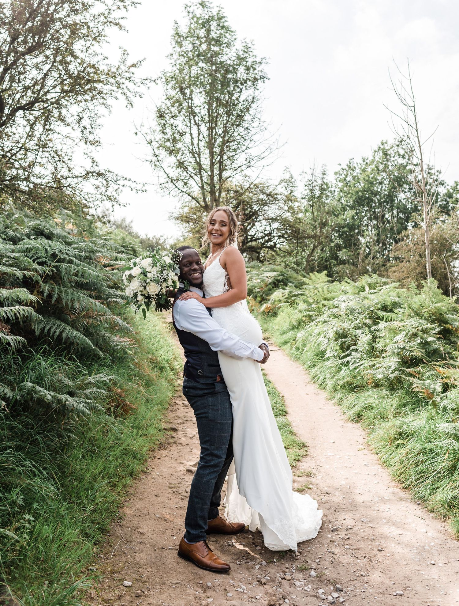 A couple embraces on a garden path surrounded by ferns on their wedding day.