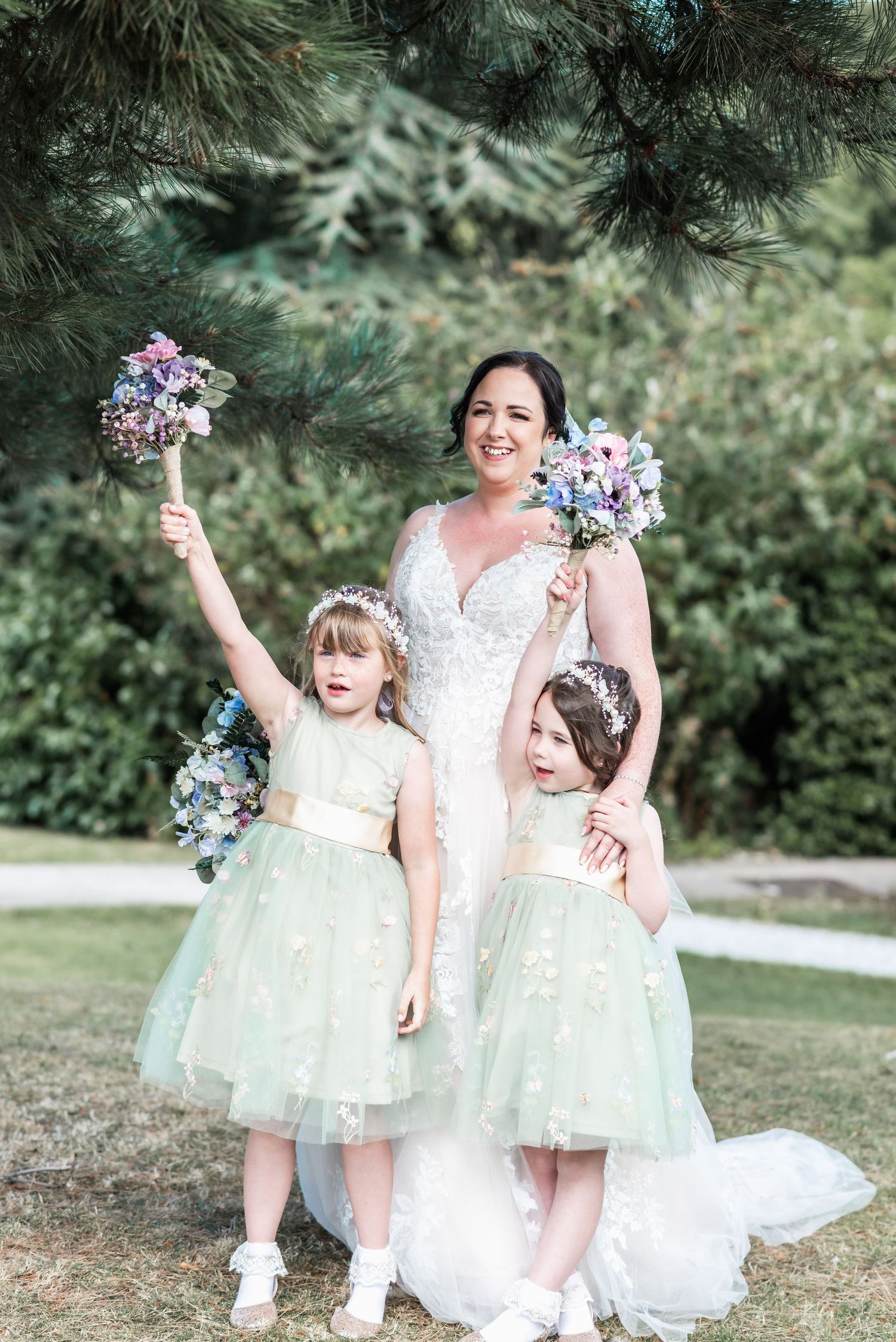 Three people in mint green and white dresses hold matching purple bouquets in garden setting.