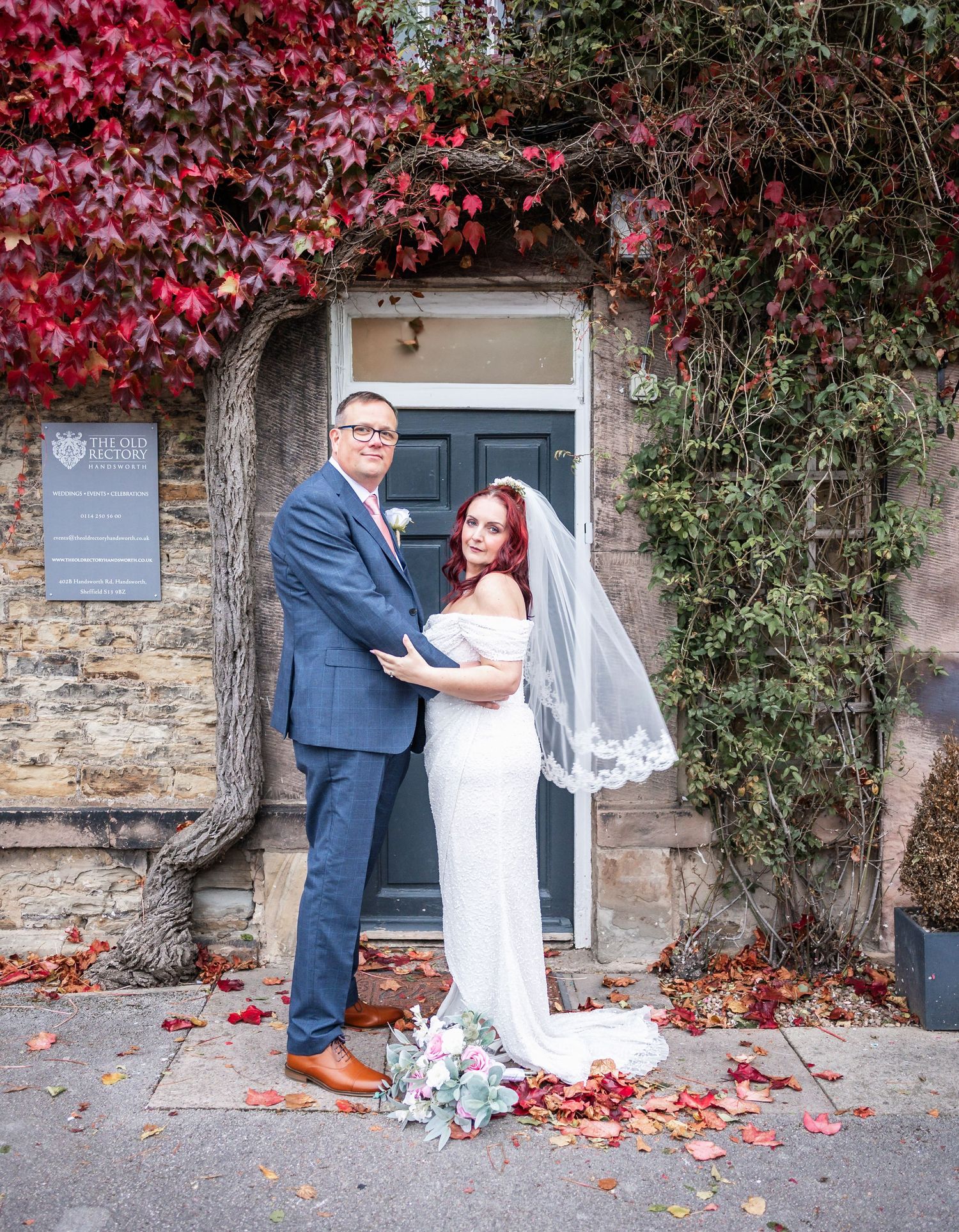 Newlyweds pose in front of a stone building entrance with red ivy and autumn leaves.