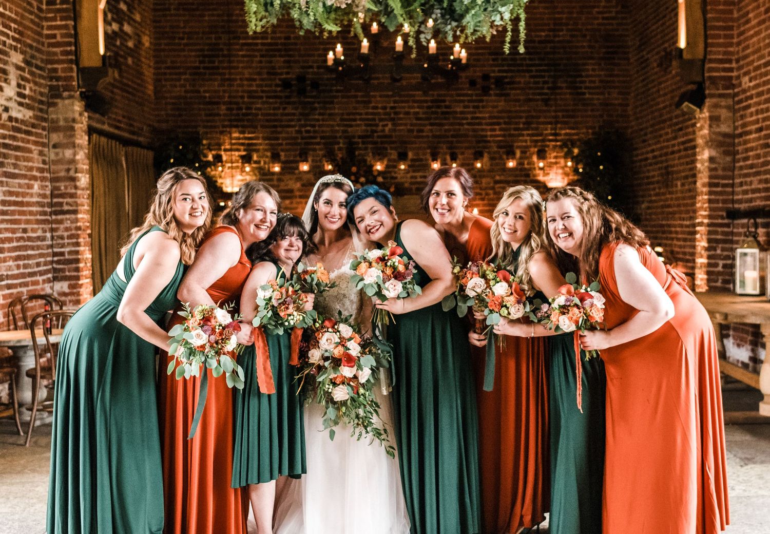 Bridal party laughing together under rustic brick arch with hanging greenery.