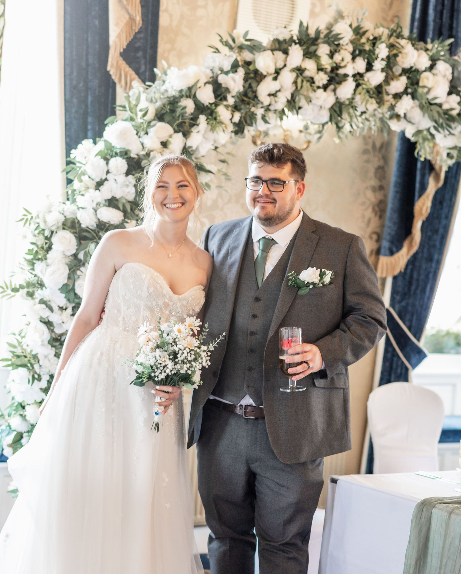 A couple poses under a floral arch at their wedding ceremony with white roses and greenery.