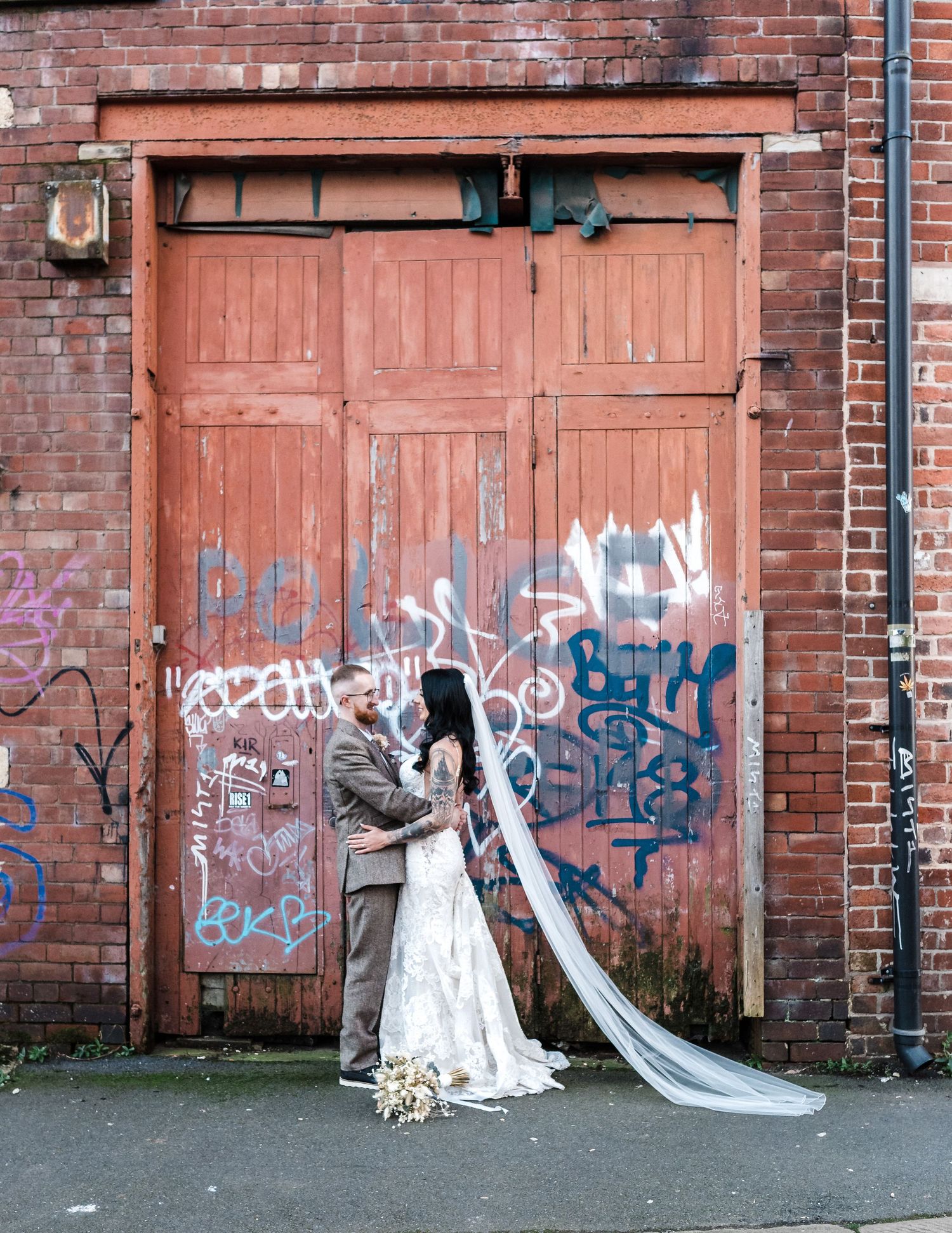 Bride poses in front of an industrial red door with graffiti while holding a bouquet.