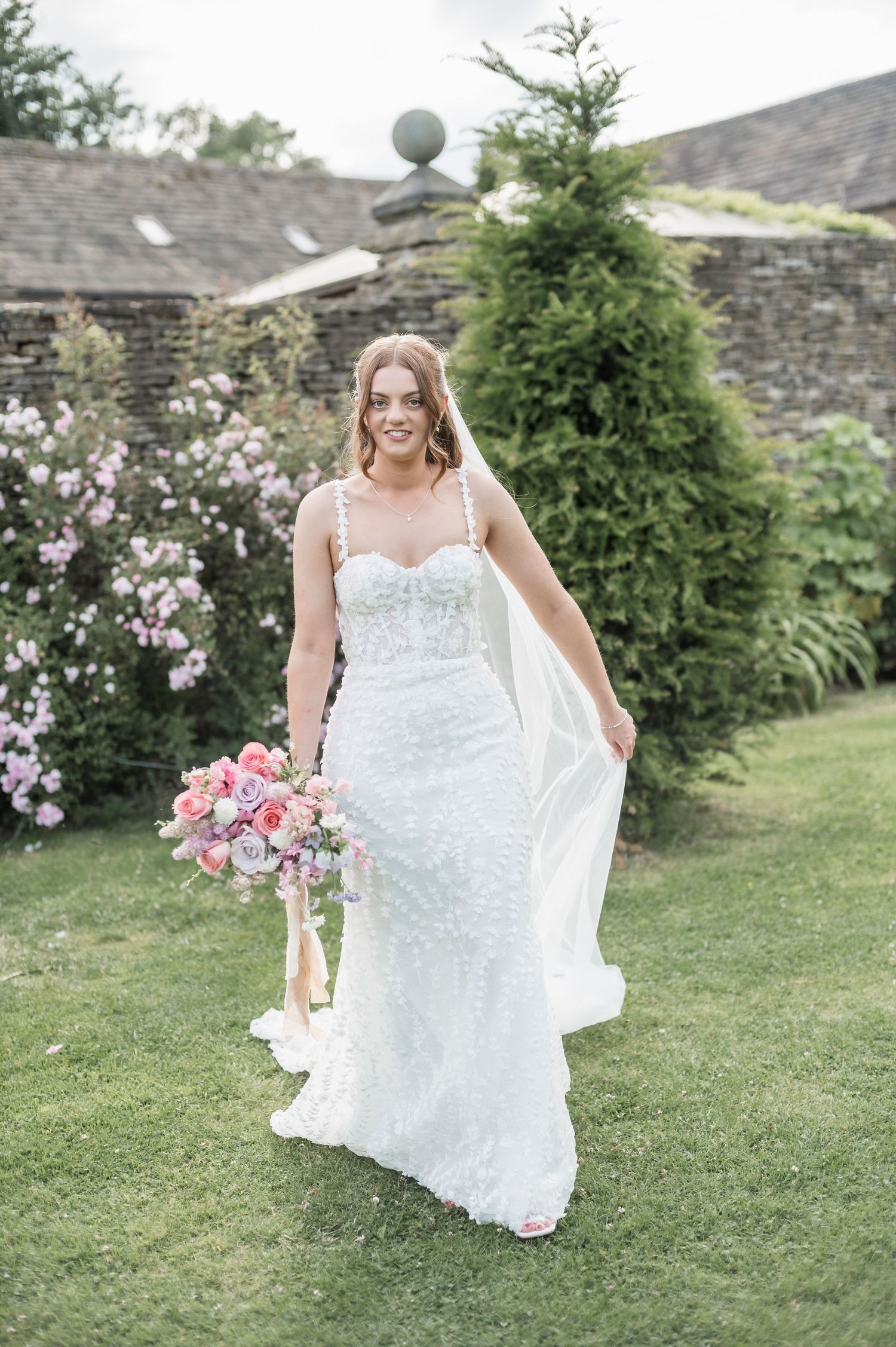 Bride in lace wedding gown holds pink bouquet in garden setting.