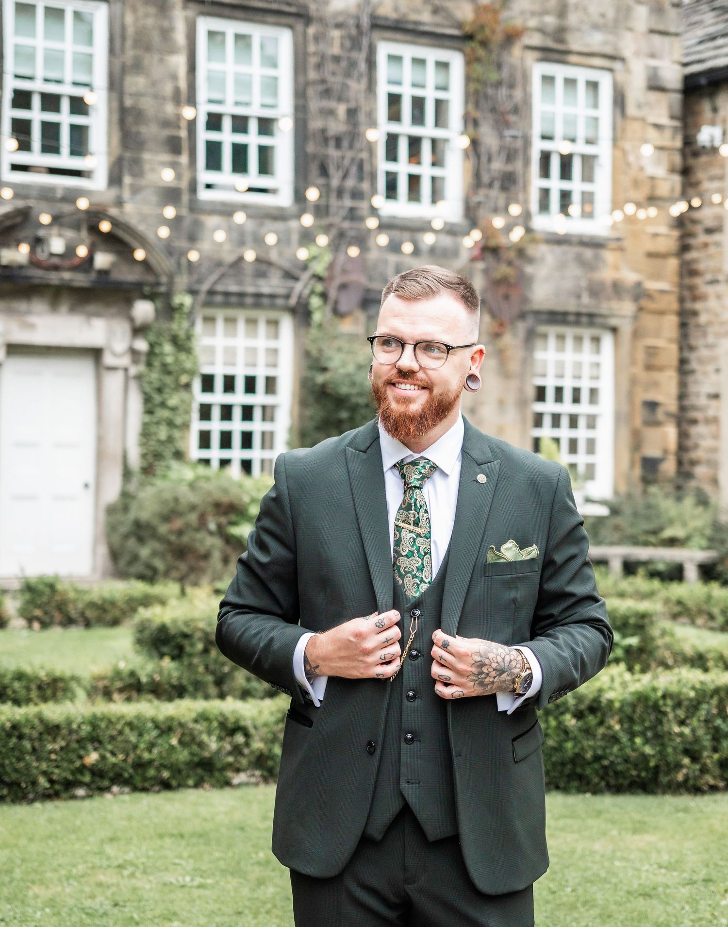 Groom in dark suit with floral tie poses in front of historic stone building with sash windows.