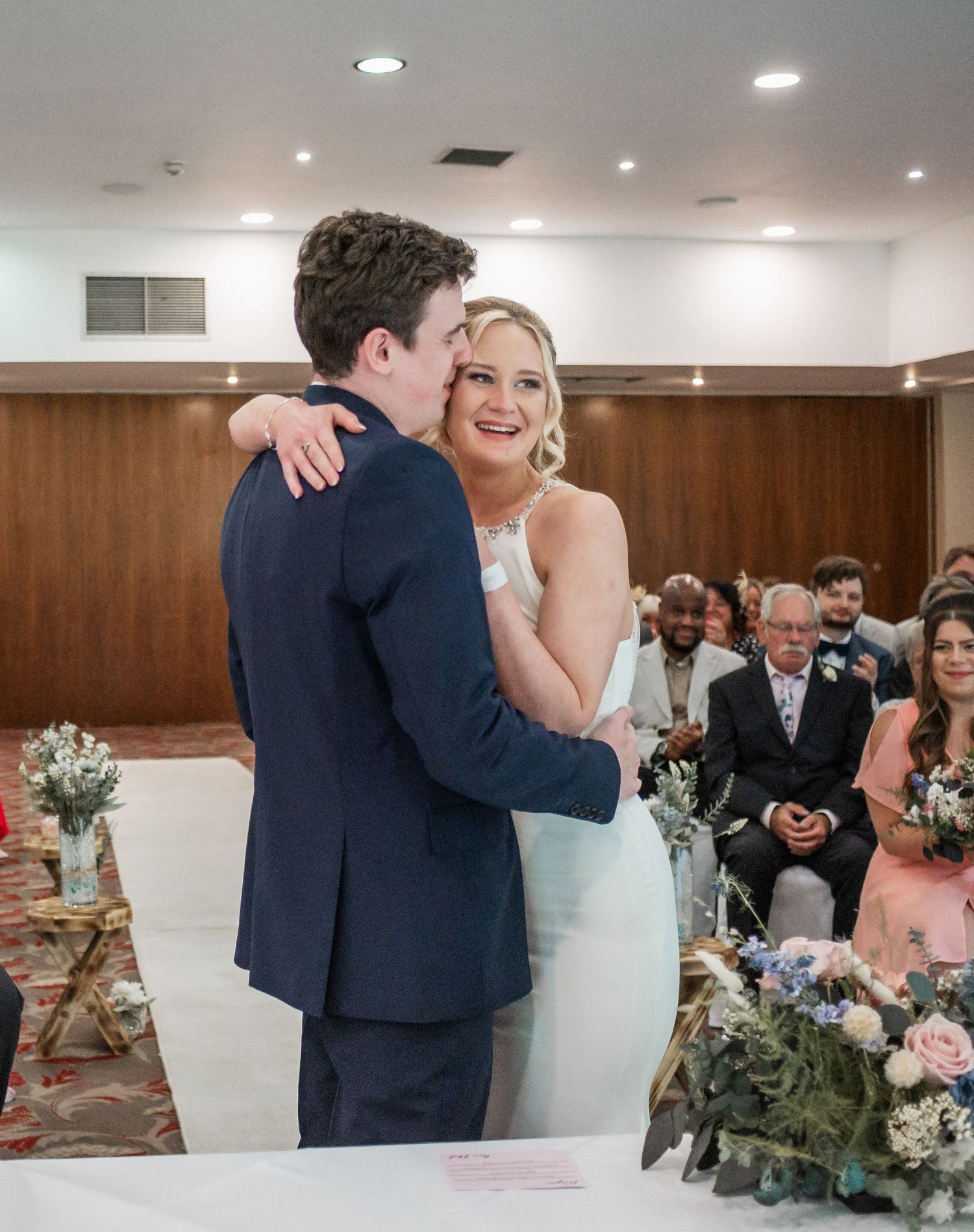 A tender moment between newlyweds at their wedding ceremony with guests looking on.