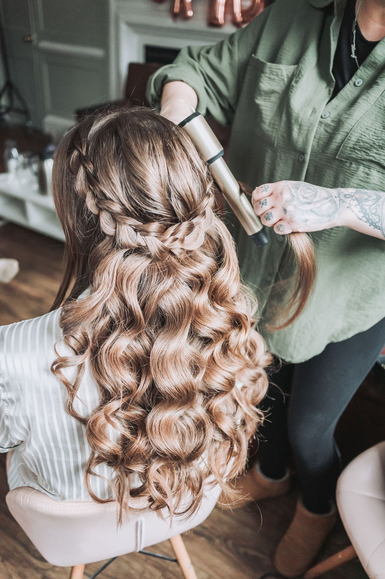 A hairstylist with tattoos creates elegant curls and braids in light brown hair for a formal occasion.