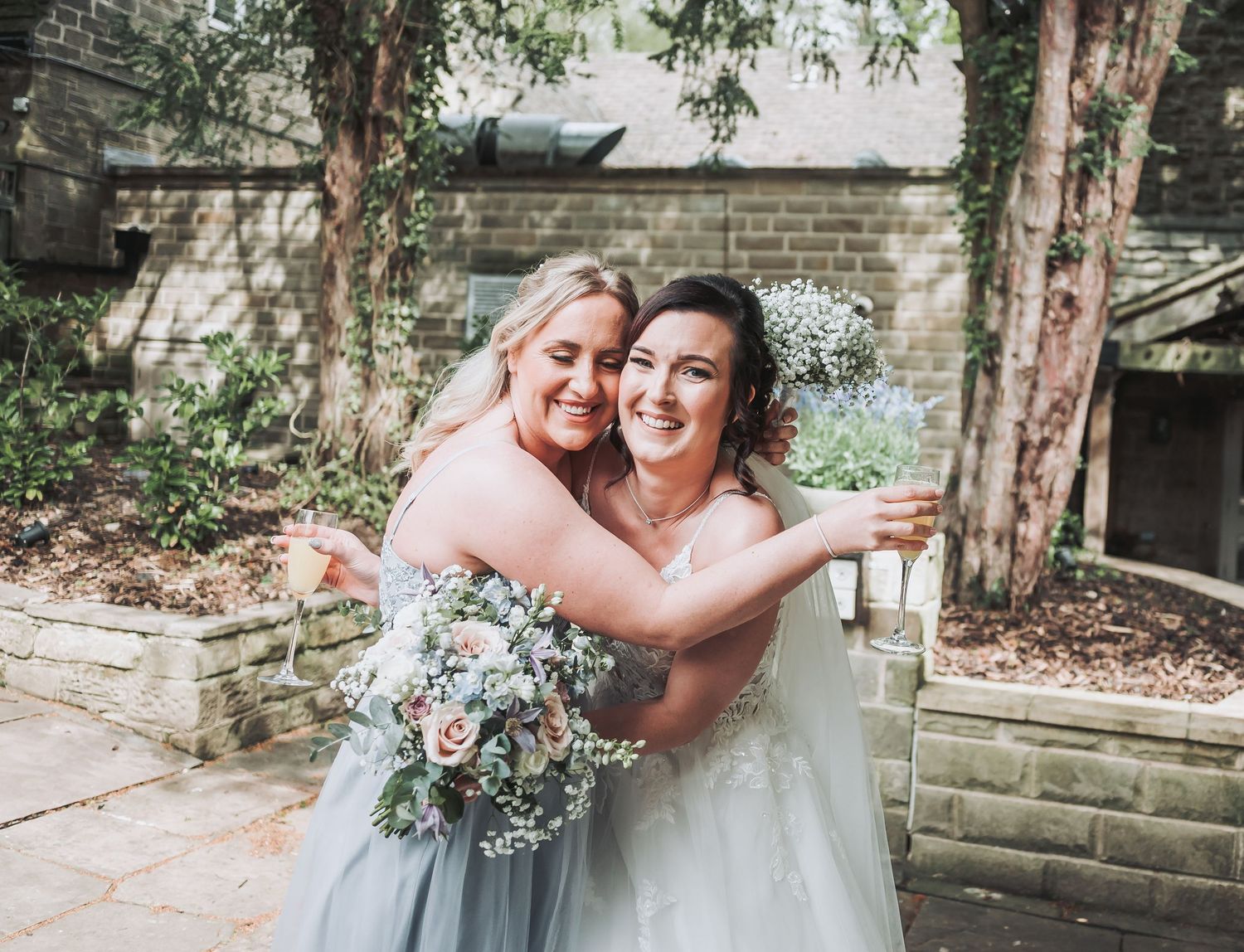 Two wedding guests hugging and smiling while holding a floral bouquet outdoors.