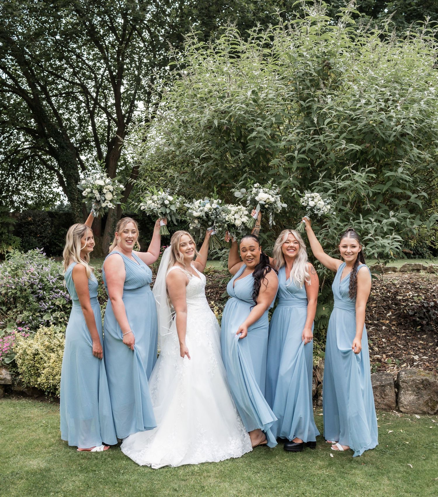 Bridal party in light blue gowns poses joyfully in garden setting.
