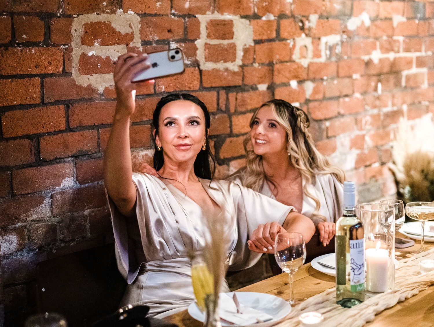 Two party guests taking selfies at a rustic brick-walled venue with drinks on the table.