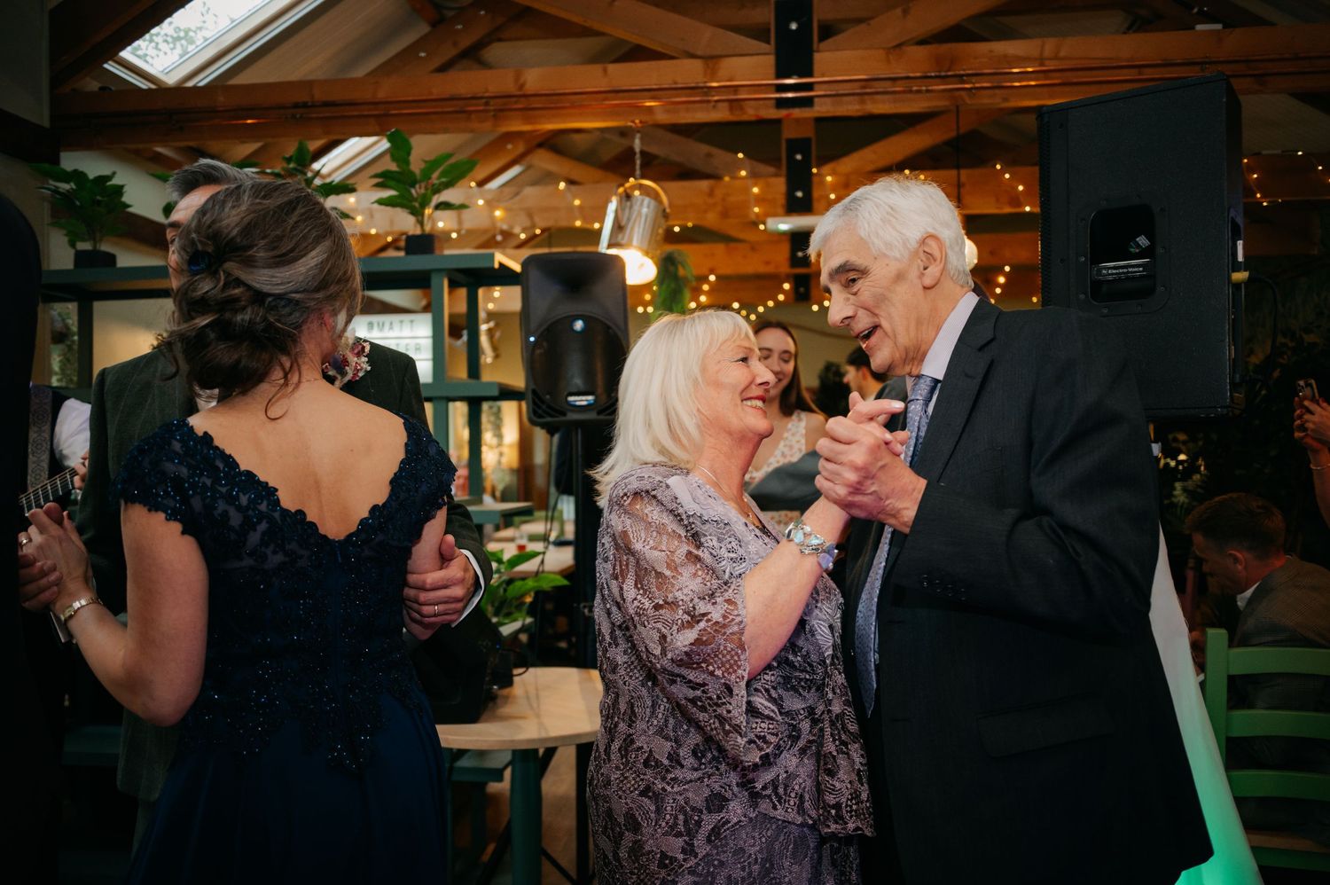 Wedding reception guests mingle in a warmly lit indoor venue with exposed beams.