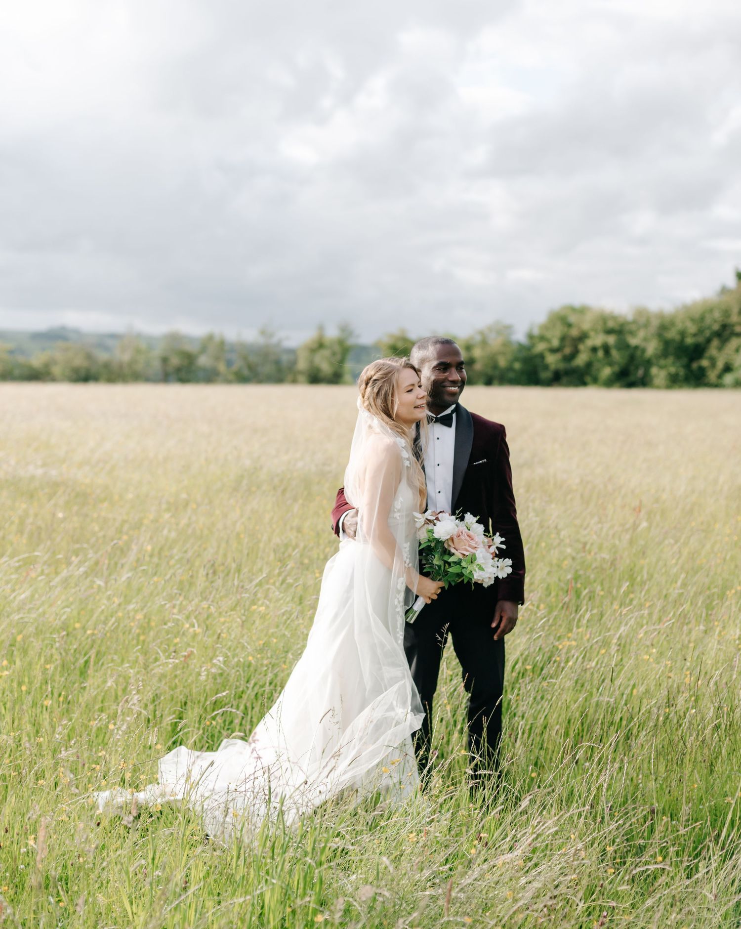 A bride and groom embrace in a lush green field, she in a white gown with train, he in a black tuxedo.