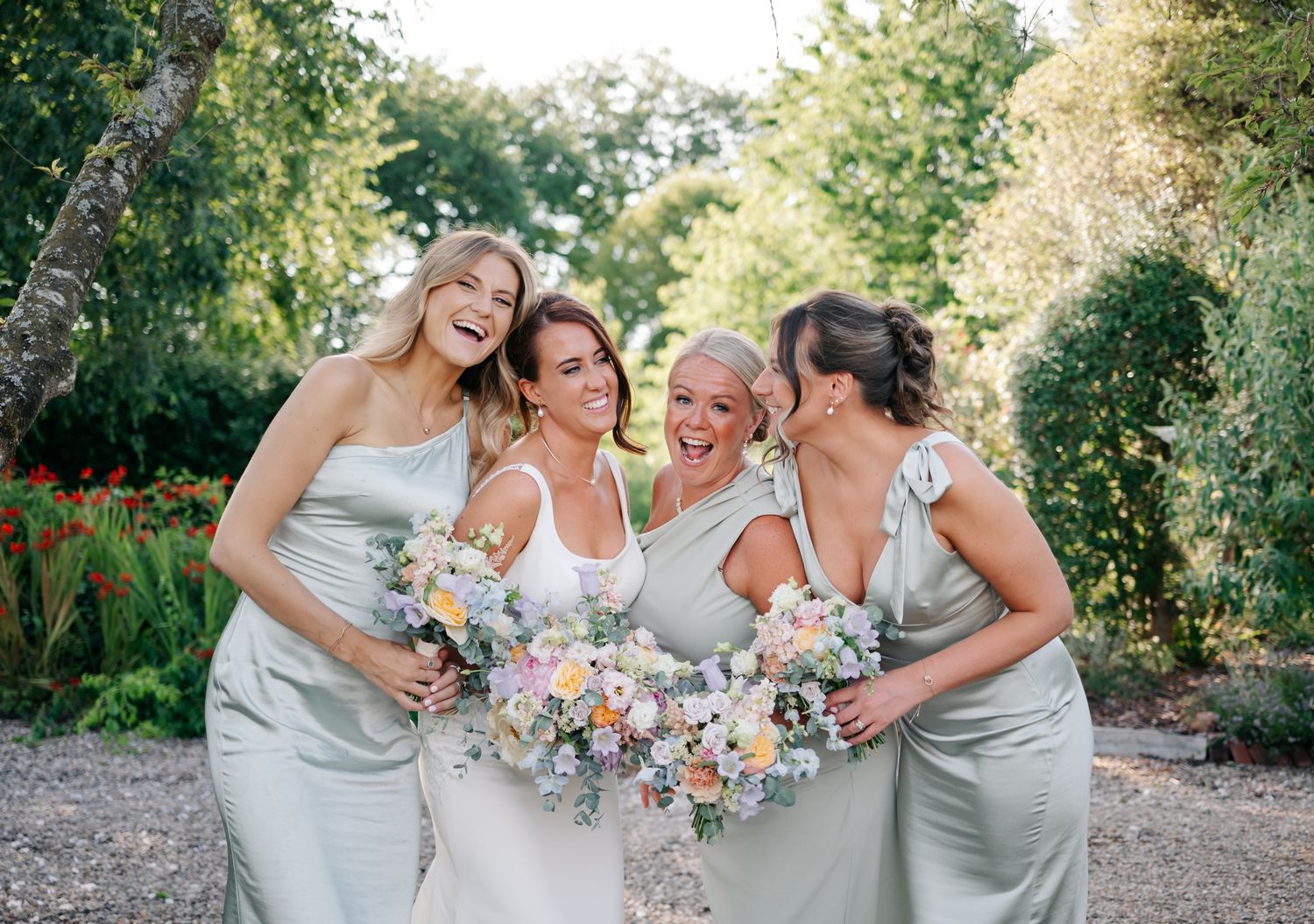 Wedding party in light gray dresses share joyful moments together outdoors on a sunny day.