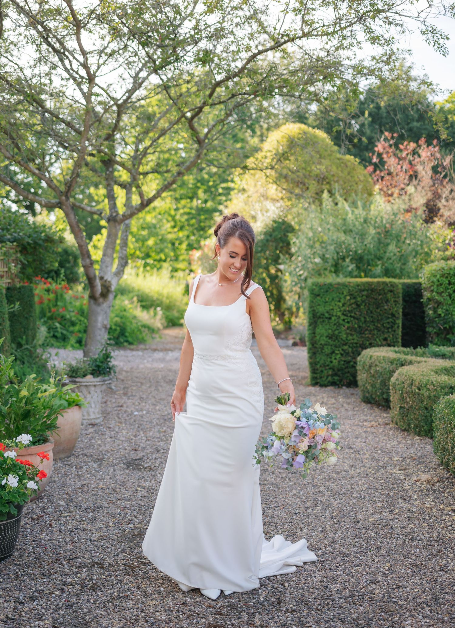 A person in a white wedding dress walks along a gravel path surrounded by manicured gardens and trees.