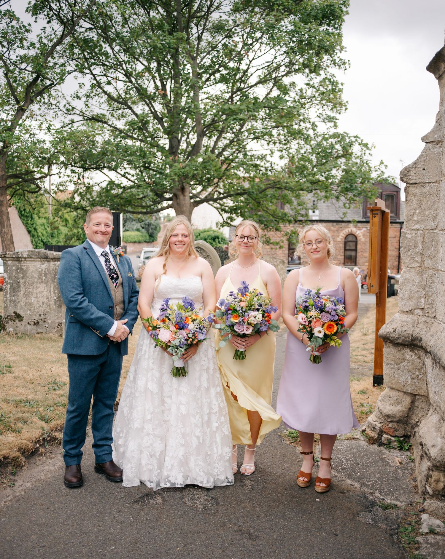 Wedding party posing outdoors against stone wall with colorful wildflower bouquets and lavender bridesmaid dresses.
