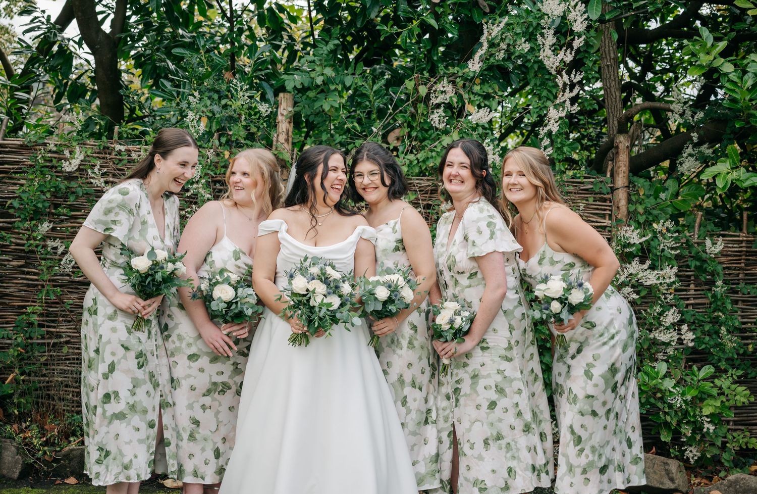 Bridesmaids in floral print dresses pose with bouquets against a lush green garden backdrop.