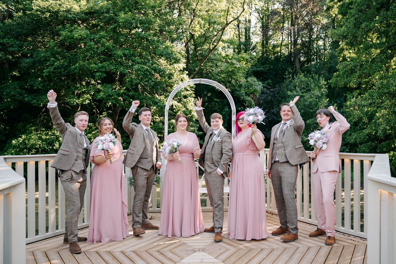 A wedding party in pink and grey celebrates on a deck with an arch and lush green trees.